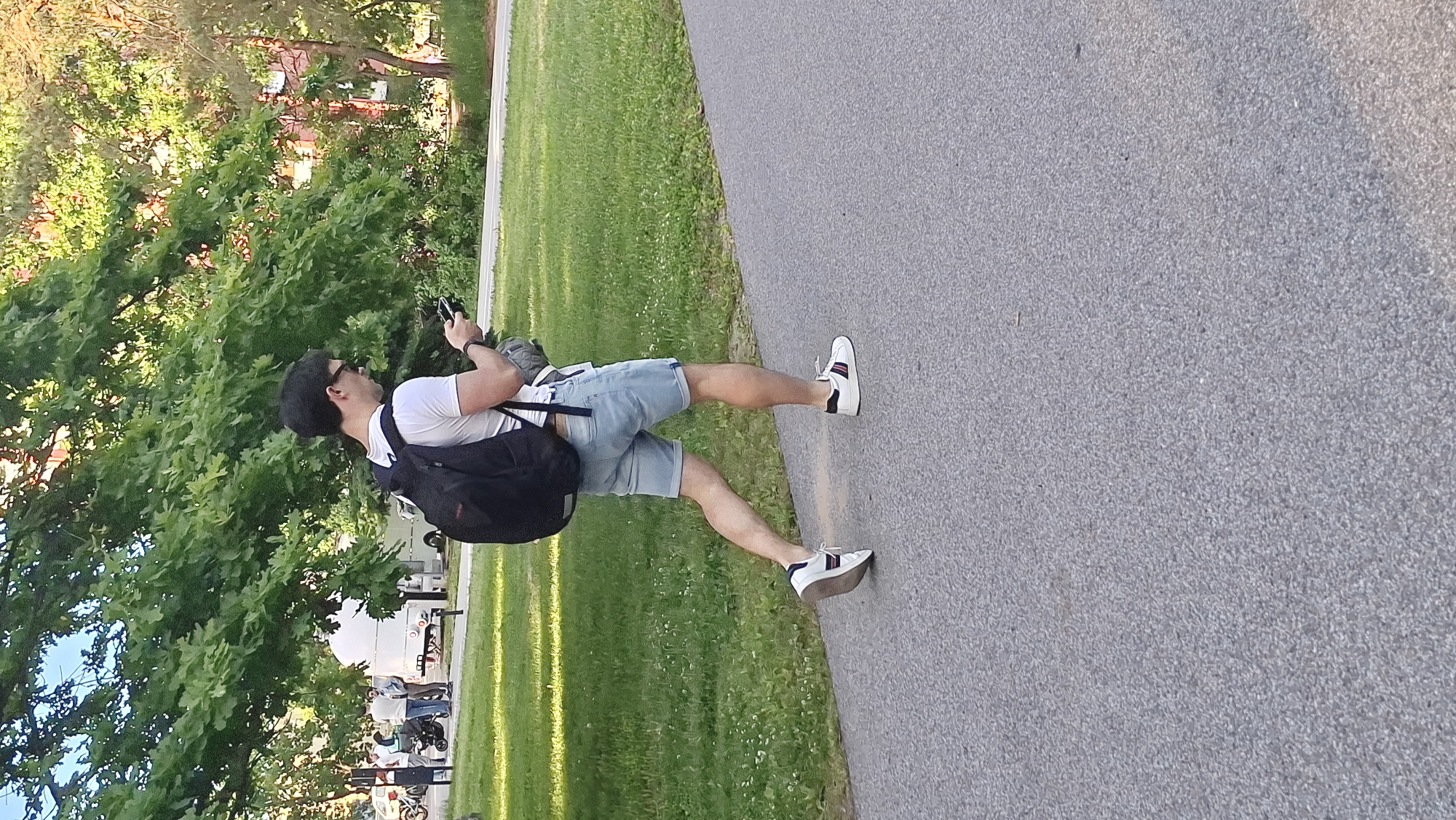 A young man walks on a paved path in a park, carrying a black backpack and looking at his phone. He is wearing a white t-shirt, light blue denim shorts, and white sneakers with red and blue stripes. The path is bordered by a grassy area with scattered white flowers, and in the background, there are trees with green foliage and a building. To the left, in the distance, several people and a white vehicle are visible, suggesting a public park setting in Tallinn, Estonia.