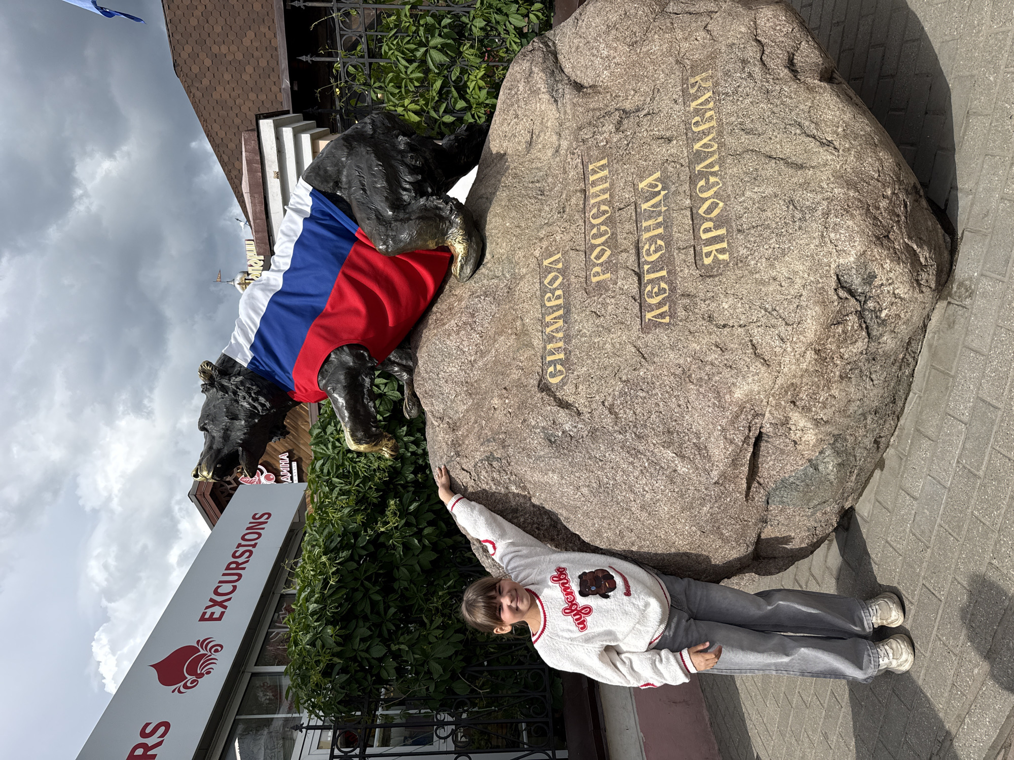 A young girl poses next to a large stone monument and a bear statue draped in the Russian flag. The monument features Cyrillic inscriptions in gold lettering, which translate to "Symbol of Russia, Legend of Yaroslavl." The scene is located in Rusanovo, Russia, as indicated by the Cyrillic text. The girl is wearing a white sweater and gray pants, and she has one hand resting on the stone. A building with an "Excursions" sign is visible in the background, suggesting a tourist area. The weather appears partly cloudy. The scene is shot during the daytime.