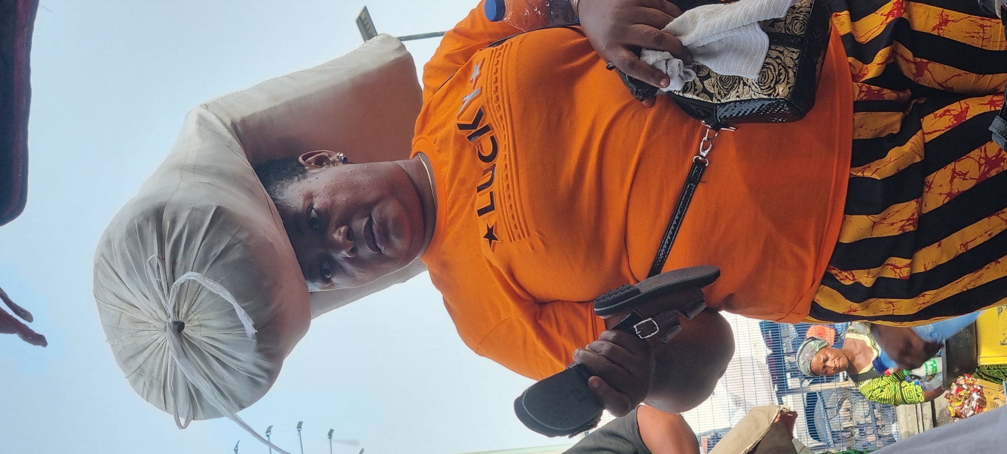 A woman in an orange shirt with "LUCKY" printed on it and a patterned skirt carries a large bag on her head. She is also holding a pair of sandals in her hand. The scene is a busy outdoor market in Lagos, Nigeria, based on the presence of other people, market stalls, and the overall atmosphere. Another woman is partially visible behind a metal barrier.