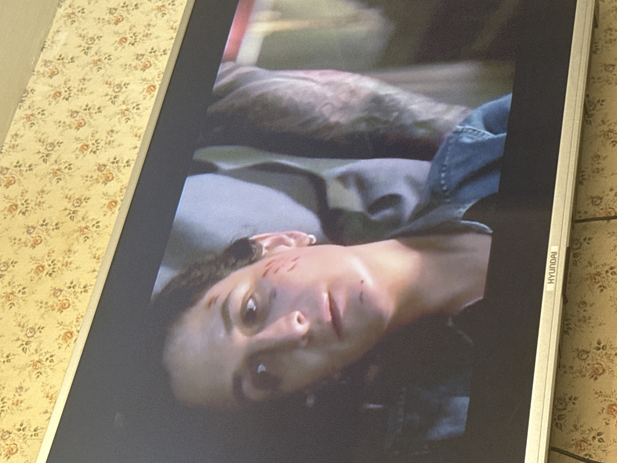 A close-up of a monitor displaying a woman with scratches on her face. The monitor is a Hyundai. The background suggests a room with floral patterned wallpaper. There is no indication of this being in Mytishchi, Russia.
