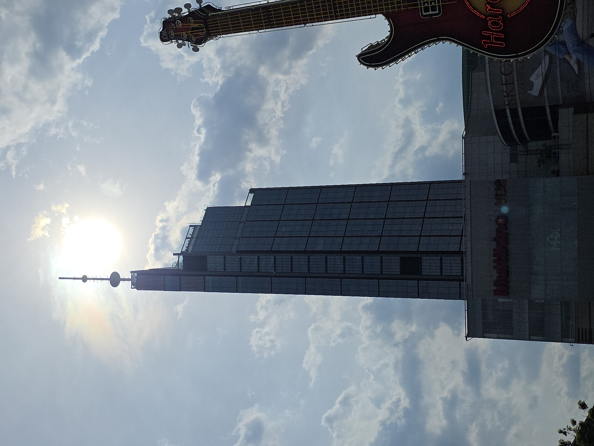 The image shows a low-angle shot of a tall building with an antenna on top, likely in Warsaw, Poland. The building has a dark, rectangular facade with a grid-like pattern. To the right of the building, there is a Hard Rock Cafe sign with a large guitar. The sky is blue with white clouds, and the sun is visible behind the building, creating a glare. The image is rotated 90 degrees counter-clockwise. The building is the Marriott hotel, and the Hard Rock Cafe is adjacent to it.