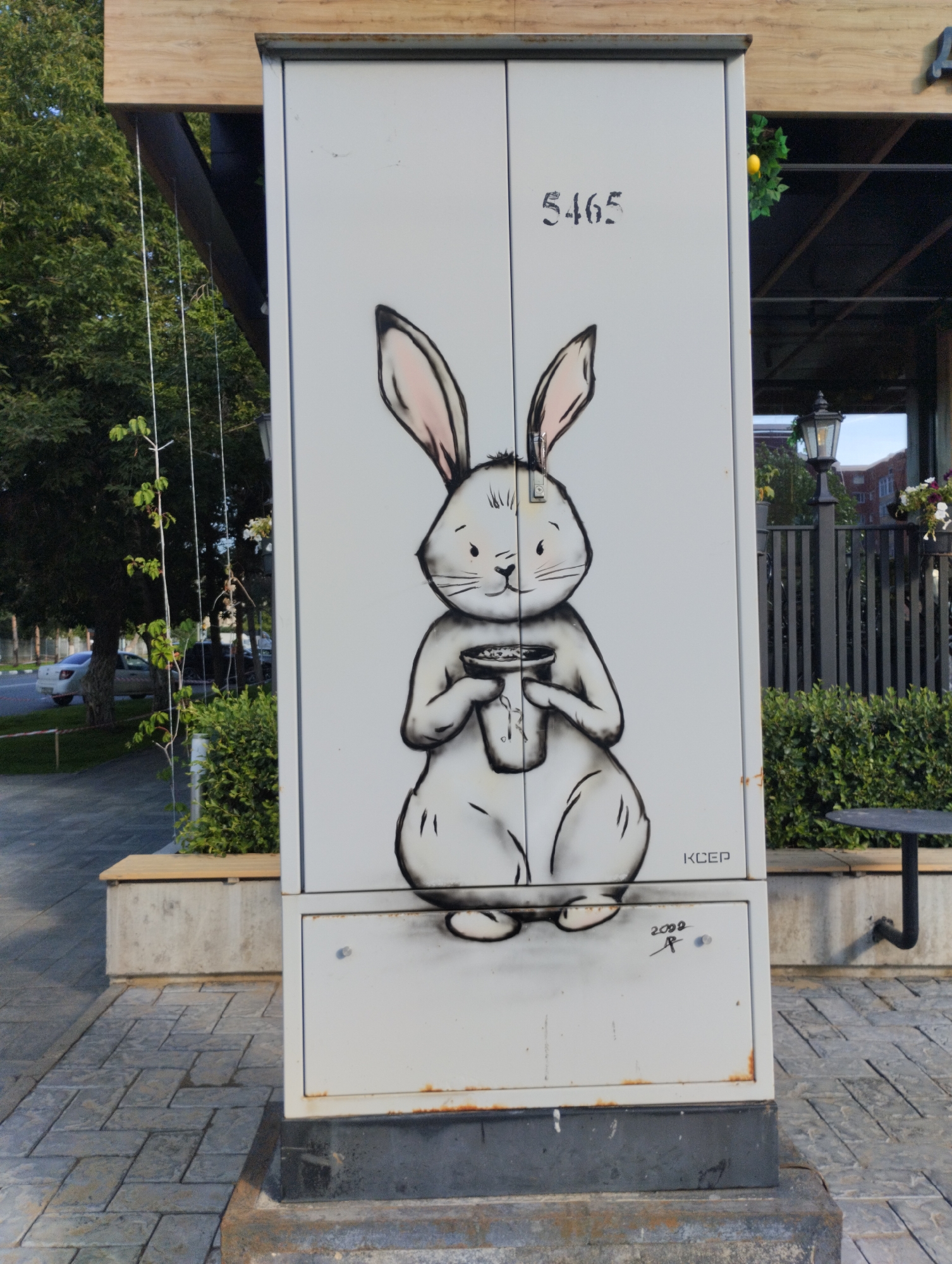The image shows a street art mural of a cartoon rabbit holding a cup, spray-painted on the side of a utility box located in Kostanay, Kazakhstan. The utility box is light gray and has the number "5465" stenciled in black above the rabbit. The rabbit is depicted in shades of gray and pink, with a white body and pink inner ears. It has large eyes and a small nose. The cup it is holding appears to contain a beverage. Below the rabbit, the artist's signature "KCEP" and the year "2000" are visible. The utility box is situated on a paved walkway. In the background, there is lush green foliage, trees, a street lamp, and a metal fence. A small circular table with a black leg is partially visible to the right of the utility box. The overall environment suggests an outdoor public space, possibly a park or a pedestrian area within the city.