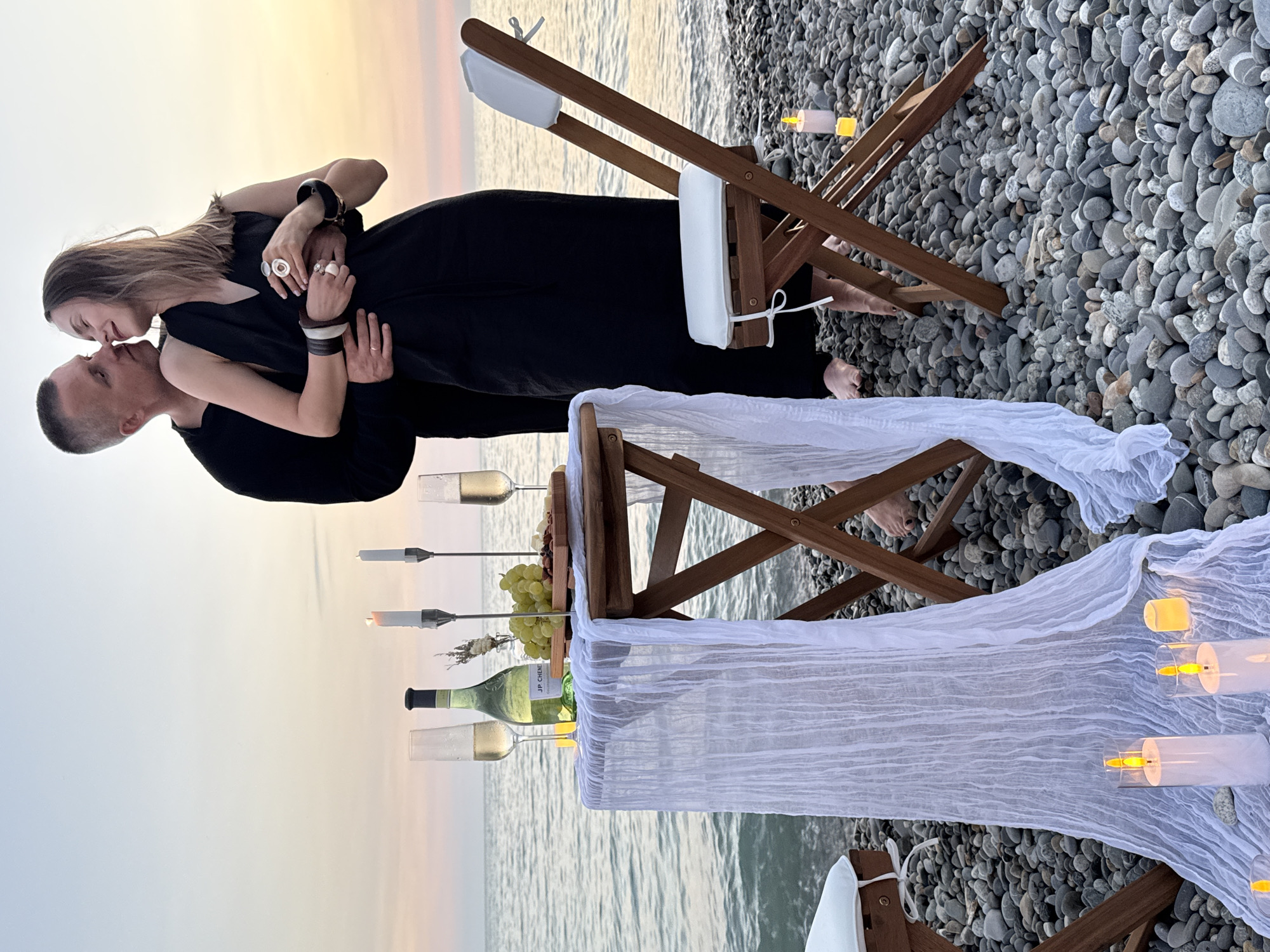 On a pebble beach in Sochi, Russia, a couple is enjoying a romantic evening by the Black Sea. The setting sun casts a warm glow on the horizon, illuminating the gentle waves. A small wooden table is set for two, adorned with a white lace runner, two champagne glasses, a bottle of white wine, a cheese platter with grapes and nuts, and two candlesticks. Two wooden chairs with white cushions are placed at the table. The couple, dressed in black, are embracing, sharing a kiss. The man has his arm around the woman, who rests her head on his shoulder. The atmosphere is intimate and serene, a perfect moment captured against the backdrop of Sochi's natural beauty.