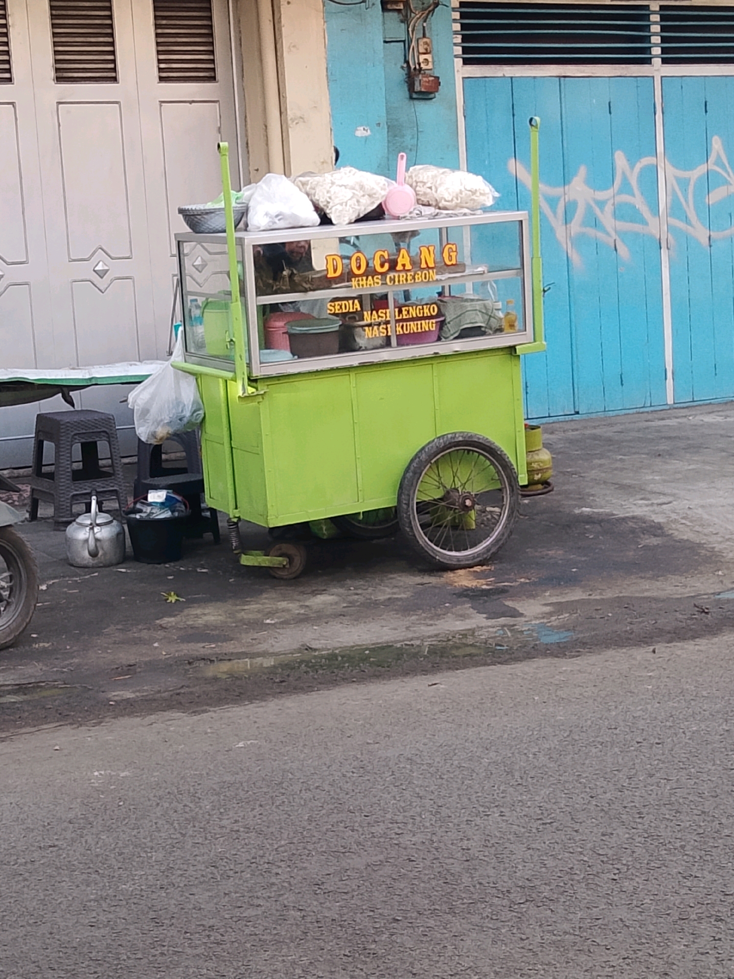 The image depicts a food cart on a street in Cirebon, Indonesia. The cart is bright green and has a glass display case filled with food items. A sign on the cart reads "DOCANG KHAS CIREBON" and lists "NASI LENGKO" and "NASI KUNING" as available dishes. In the background, a blue door with white graffiti and a beige building with large doors are visible. A gas cylinder is located behind the cart. On the left side of the image, there are two black stools, a metal teapot, and a black bucket. The ground is paved and appears wet in some areas. The overall scene suggests a street-side food vendor operating in a typically urban environment within Cirebon.