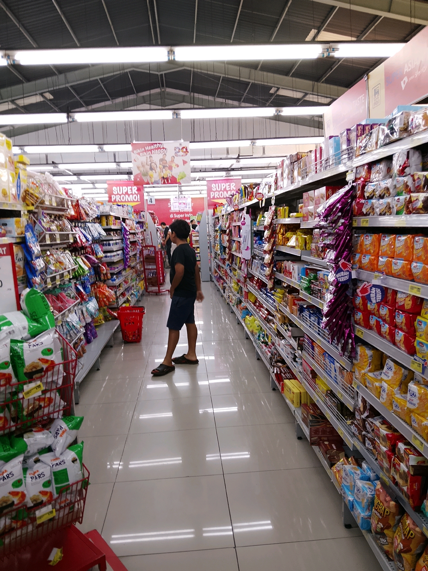 The image depicts the interior of a supermarket aisle in Malang, Indonesia. The aisle is lined with shelves stocked with various packaged food items, predominantly snacks and biscuits. Prominent signage indicates "SUPER PROMO!" in red banners hanging from the ceiling, suggesting a sale or promotional event.
A young person, appearing to be a male teenager, is standing in the aisle, facing away from the camera and looking towards the shelves. He is wearing a dark t-shirt and blue shorts, with black sandals. He is standing near a bright red shopping basket placed on the floor.
The supermarket environment is characterized by a clean, tiled floor reflecting the overhead fluorescent lighting. The ceiling is a metal structure with exposed pipes and lighting fixtures. The overall impression is of a well-lit and organized retail space. The presence of "SUPER PROMO!" banners and the abundance of packaged goods suggest a typical grocery shopping experience.