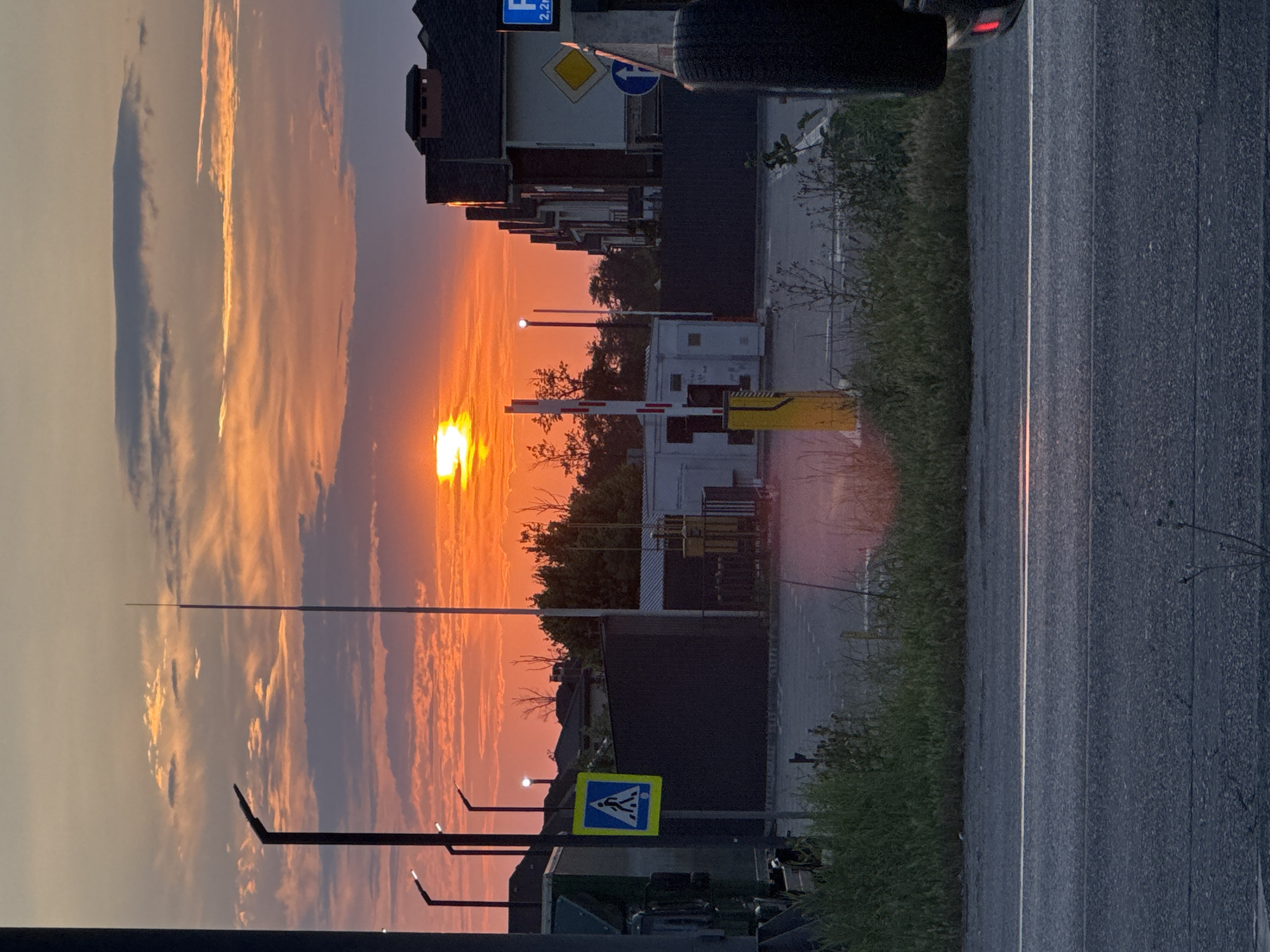 The image depicts a sunset scene in a suburban or industrial area, likely on the outskirts of a city in Russia. The sun is low on the horizon, casting a vibrant orange and pink glow across the sky, illuminating scattered clouds. In the foreground, a paved road with a white line marking the lane is visible. Beyond the road, a grassy area transitions to a fenced-off area with buildings, streetlights, and a traffic barrier.
Several key objects are present:
- **Traffic Lights/Barriers:** A red and white striped traffic barrier stands prominently in the middle ground, suggesting a checkpoint or entrance. A yellow bollard is also visible near it.
- **Streetlights:** Modern, minimalist streetlights with angular designs are spaced along the left side of the image.
- **Pedestrian Crossing Sign:** A blue square sign with a white pedestrian symbol is mounted on a pole, indicating a crosswalk.
- **Vehicles:** A large, dark-colored vehicle, possibly a truck or utility vehicle, is partially visible on the far left. The rear tire of another vehicle is visible on the right side of the image, with a red taillight illuminated.
- **Buildings:** Rows of modern houses with dark roofs are visible on the right side of the image. In the center, a low-rise building with white walls and some industrial-looking equipment is present.
- **Vegetation:** Grassy verges line the road and the perimeter of the fenced area. Some trees are visible in the background.
- **Sky:** The sky is dramatic with the setting sun and colorful clouds.
The scene appears to be captured during the late evening. The overall atmosphere is quiet, with no people visible in the frame. The image is taken from a slightly elevated perspective, looking across the road towards the buildings and the sunset.