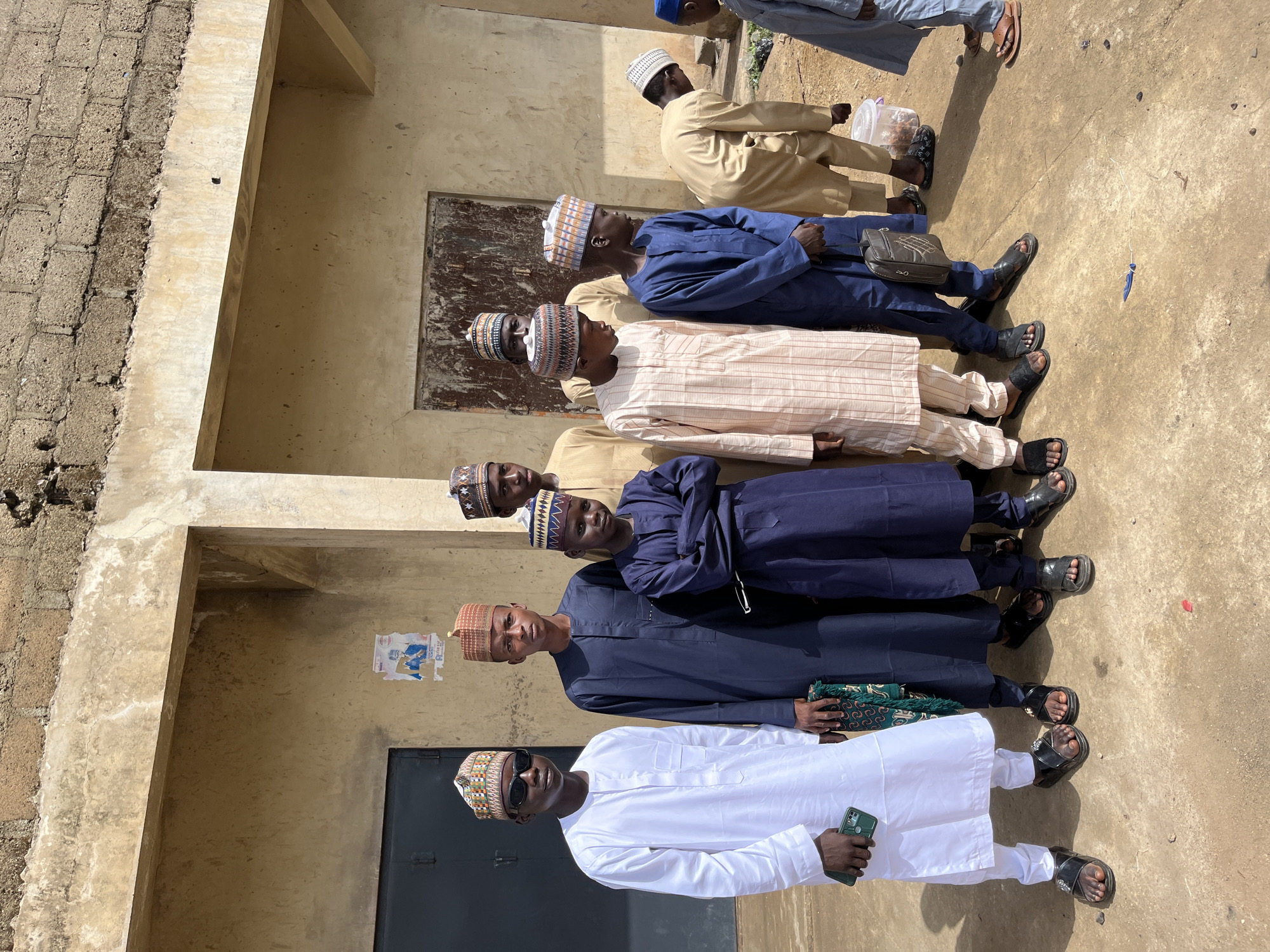 In Naraguta Hausawa, Nigeria, a group of men stands in a line outdoors next to a building with a weathered facade. The men are wearing various traditional attire including embroidered caps, robes in shades of white, beige, and blue. Many are wearing sandals. One man in the front is wearing sunglasses and holding a cell phone. A few posters or signs are affixed to the building wall.