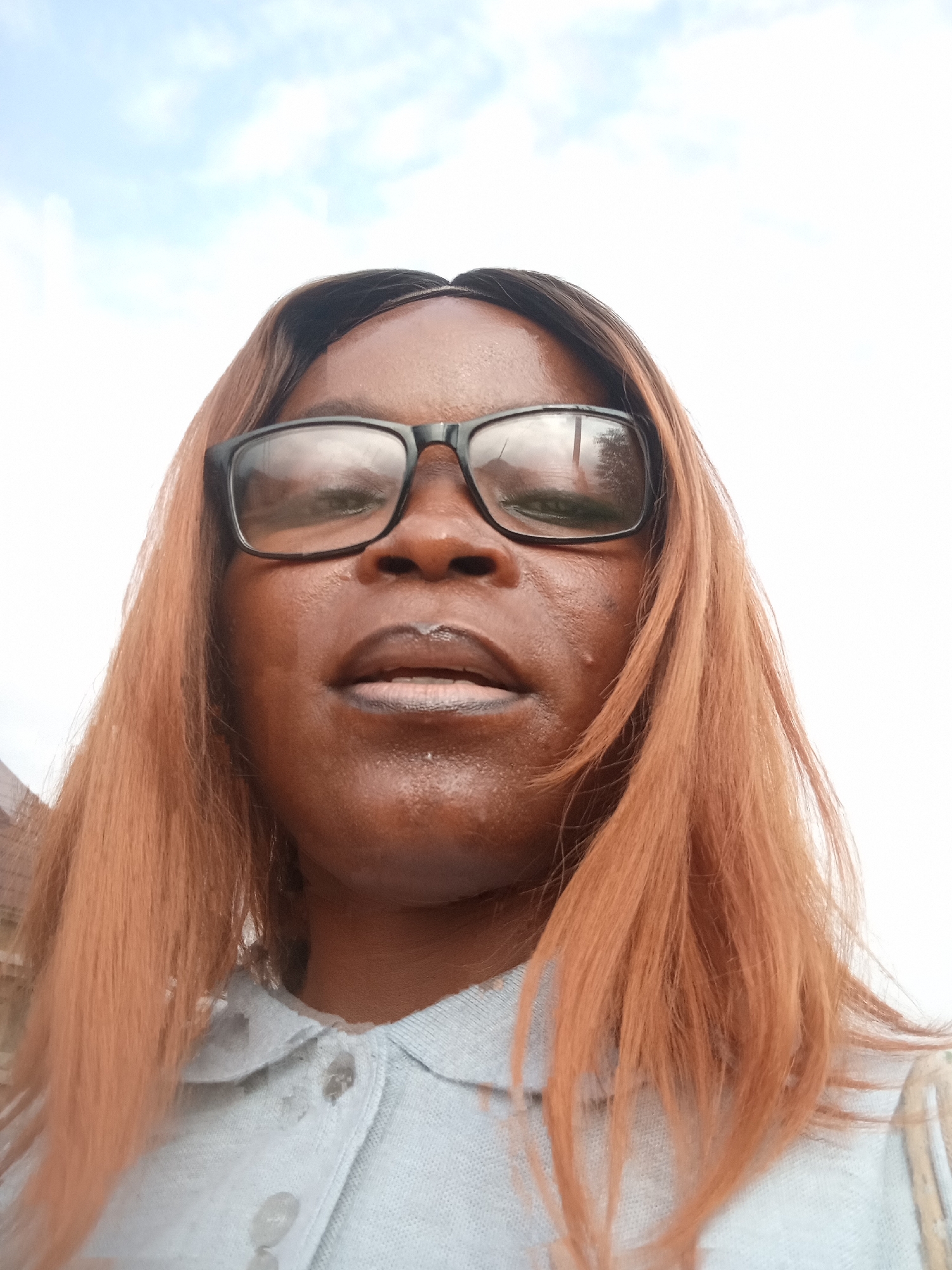 A woman with medium-toned skin, wearing black-rimmed glasses and a light blue collared shirt, is pictured in a close-up, low-angle selfie. Her reddish-brown hair is parted in the middle and falls around her shoulders. The background consists of a bright, overcast sky with a hint of clouds, and the suggestion of buildings and foliage at the lower left of the frame. The image appears to have been taken outdoors, possibly in an urban or semi-urban setting in Kason, Nigeria.