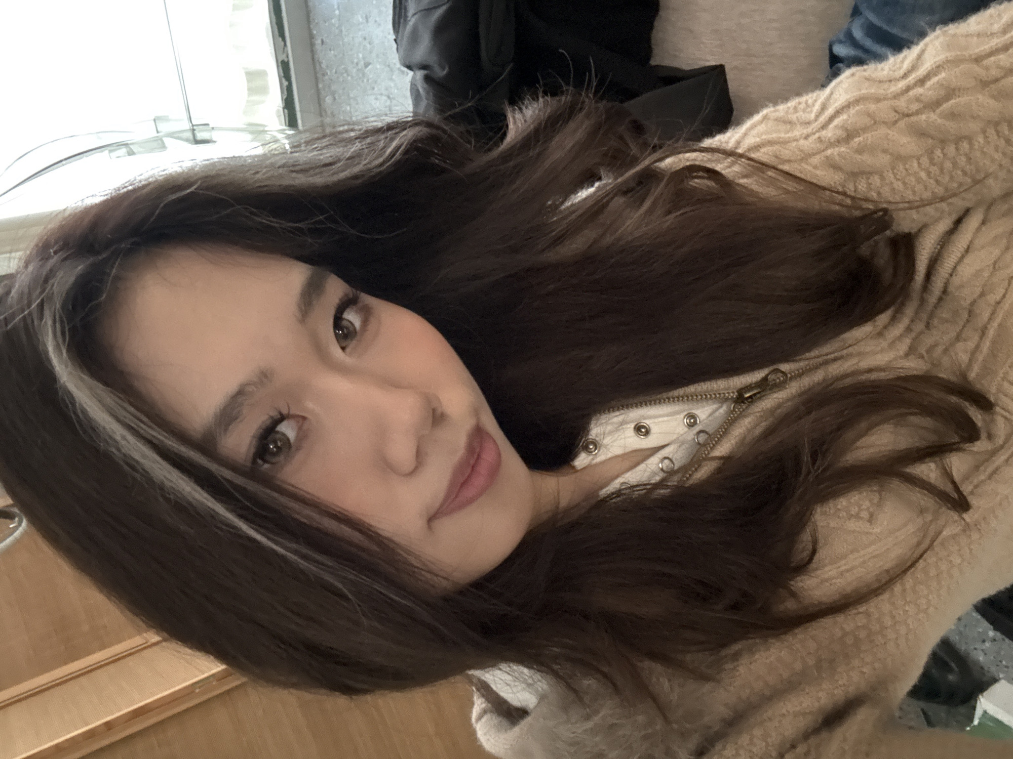 The image depicts a young East Asian female, captured indoors, stated to be in the city of Liangjiang, China. The subject, positioned centrally, has long, dark brown hair, parted to reveal a prominent light-colored streak on the viewer's left side of her head. Her brown eyes are open, with defined eyelashes, and she gazes directly at the camera with a slight, closed-mouth smile. She wears a light beige or tan cable-knit sweater or cardigan with a visible zipper closure. Beneath it, a white garment with a row of metal snap buttons is discernible at the neckline. The background elements include a light-colored wooden panel or furniture piece on the upper left. To the upper right, a window with bright daylight is visible, showing parts of a window frame. Below the window, on the right side of the frame, there are indistinct dark objects, possibly folded clothing, and a section of soft, textured fabric, with a dark blue material partially visible at the bottom right.