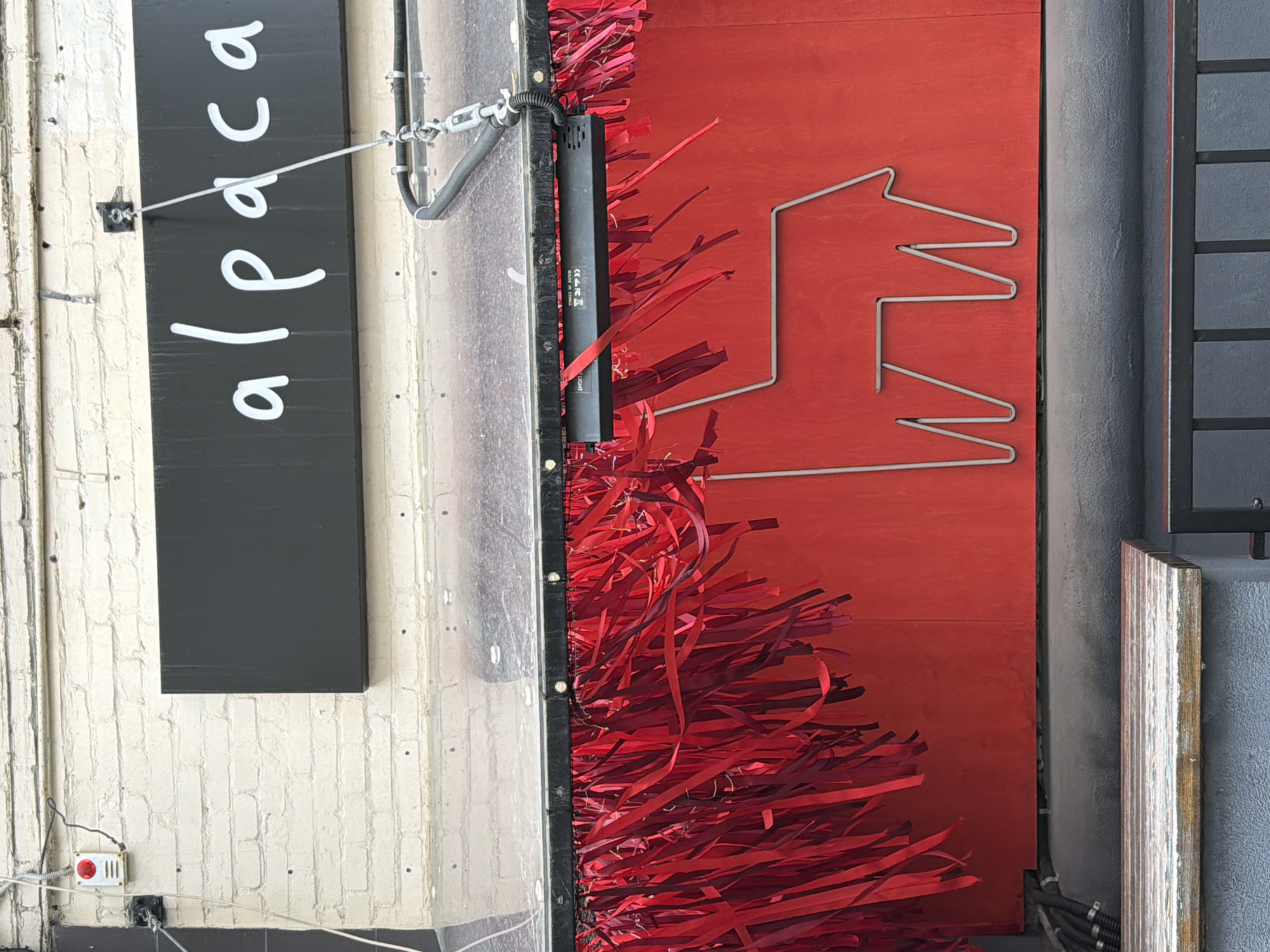 The image captures a building facade with several notable features. A black sign with the word "alpaca" written vertically in white lettering is mounted on a textured, off-white brick wall. To the right of the sign is a clear, possibly glass or acrylic, panel extending downwards. Next to this panel is a vertical black object, possibly a light fixture, adorned with red decorative streamers or ribbon. Adjacent to this is a red door or wall section bearing a minimalist, wireframe design resembling an arrow or a stylized house. The door is flanked by gray or concrete pillars. A small white device with a red button is visible on the brick wall near the bottom left corner. Based on the sign "alpaca" and the architectural style, this image might be from Moscow, Russia, as the restaurant "Alpaca" is located there.