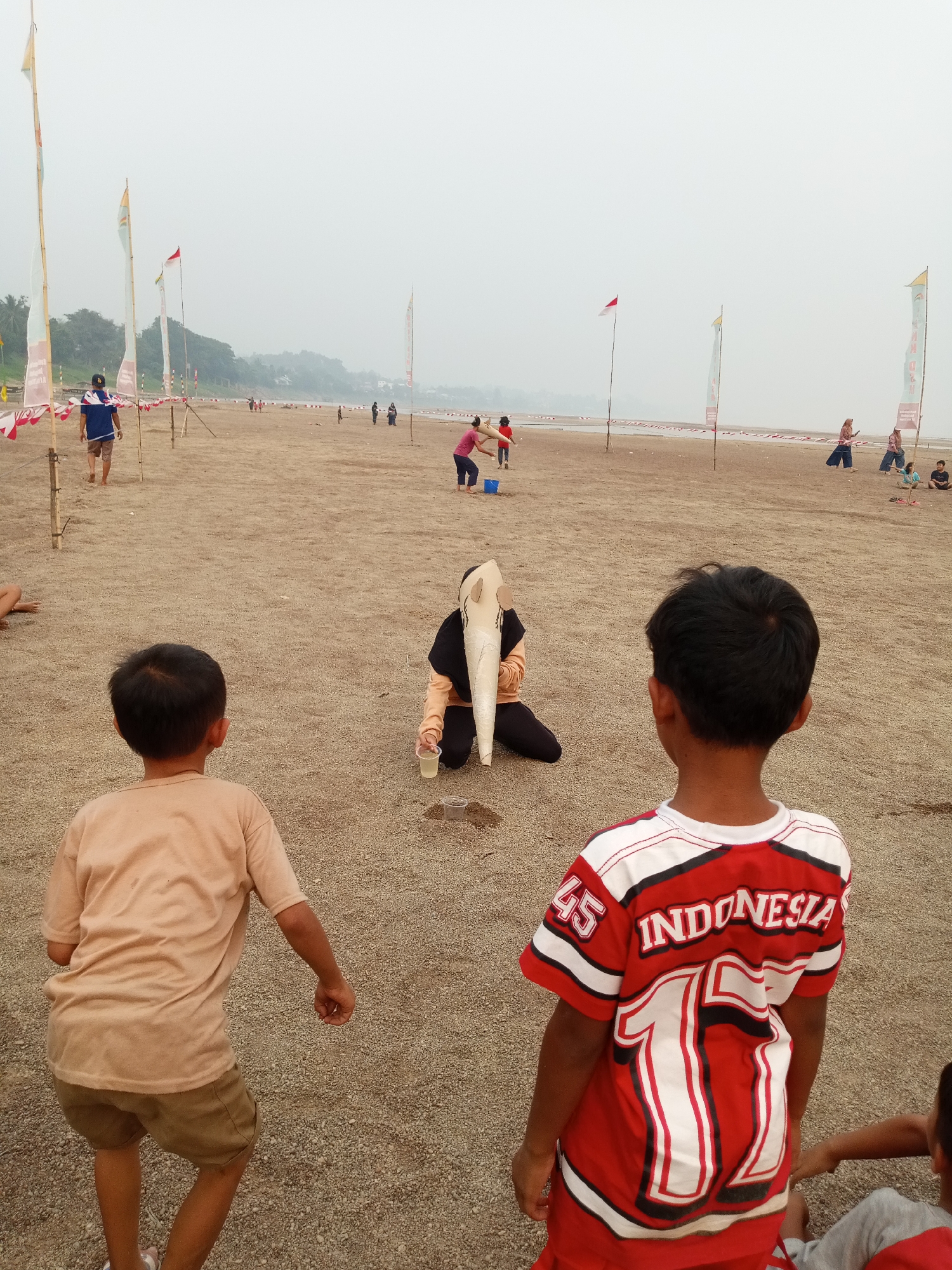 On a hazy day at a beach in Kapuas, Indonesia, several people are engaged in activities. In the foreground, two young boys are facing away from the camera. The boy on the left wears a tan t-shirt and khaki shorts. The boy on the right wears a red and white jersey with "INDONESIA" and the number "11" visible on the back, along with "45" on the sleeve.
In the middle ground, a person wearing a black hijab and an orange top is kneeling on the sand, holding a white object that resembles a cone or funnel. Next to this person are two small plastic cups, one of which appears to contain a light-colored liquid. Other individuals are scattered across the beach in the mid-distance, some walking and others appearing to be participating in a game or event.
The beach is sandy and appears to be a public area. Several tall poles with flags are planted in the sand, with some featuring red and white banners, likely indicating a celebration or event. In the background, a body of water is visible, beyond which is a treeline and a hazy sky. The overall impression is of a community gathering on the waterfront.
