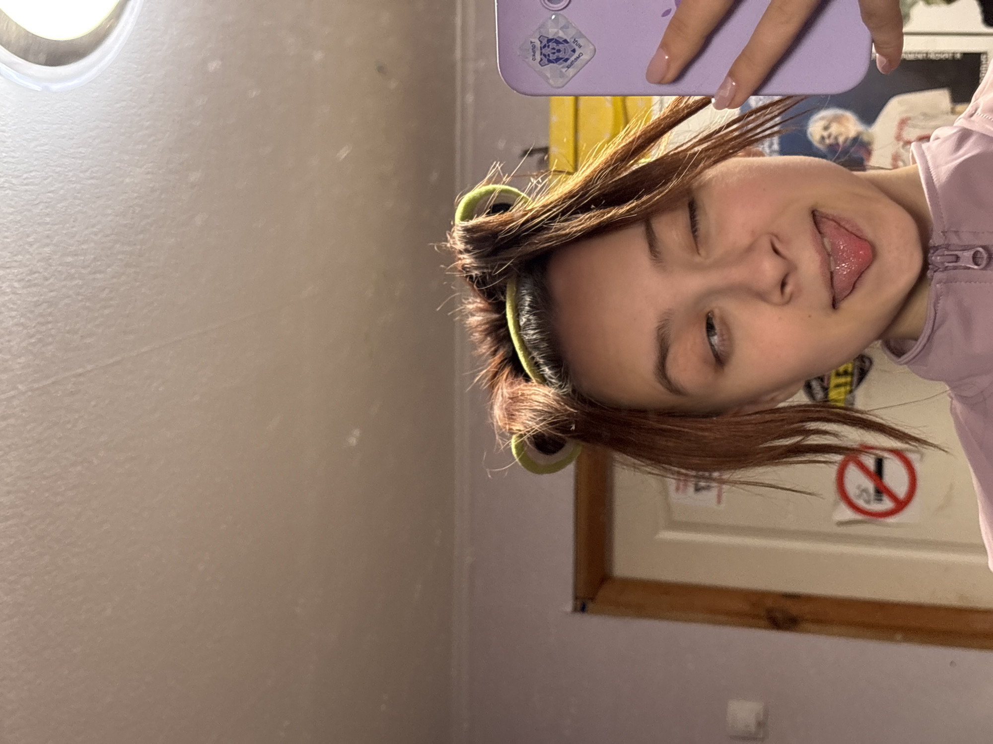 The image displays a selfie taken by a young person, presented in a portrait orientation but rotated 90 degrees counter-clockwise. The subject, likely female, has light brown hair styled with several green hair rollers visible on the sides of her head, indicating she is in the process of styling her hair. She is wearing a light purple or pink zippered top. Her left hand, adorned with light pink polished nails, holds a purple smartphone that covers the upper left portion of her face. A clear sticker featuring a geometric design, possibly a turtle or tortoise, and faint text including "OMROT" is affixed to the phone case.
The person is winking with their visible right eye and playfully sticking out their tongue, conveying a lighthearted and silly mood.
The background suggests an indoor setting, likely a personal room within a home in Прикамский, Russia. The wall behind her is a light, possibly beige or off-white color, with some texture and specks visible. In the upper left corner, a bright circular light fixture is prominent. To the right of the subject, a wooden door frame or mirror frame is visible. Further in the background, there are hints of posters or other wall decorations, including a red circular "no" symbol sign with a diagonal line through it, positioned slightly to the right of the subject's head. A small, indistinct white object lies on the floor or a surface in the lower right portion of the frame. The overall impression is a casual, personal moment captured during hair styling.