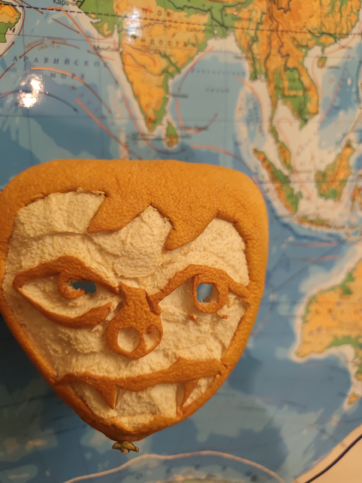 An orange peel carved into a grotesque face is positioned in front of a world map. The map displays a predominantly blue ocean area, with yellow landmasses visible, particularly in the upper right quadrant representing Asia. The carved orange peel face features exaggerated facial features including angular eyebrows, circular eyes, a circular nose, and a toothy grin with pointed incisors. The carved areas are white. The image's focus is primarily on the orange peel carving, but no identifiable information about Vlaskovo, Russia is found in this photo.