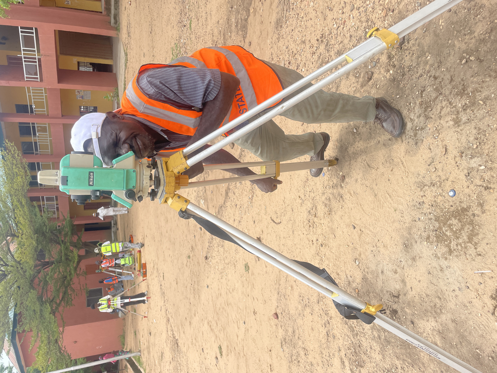 The image depicts a man in a reflective orange vest and a white cap operating a Nikon total station, a surveying instrument, in an outdoor, dusty environment. He is looking through the instrument's eyepiece and appears to be engaged in surveying work. The background shows a building with red and yellow facades, possibly a school or administrative building, and several other individuals in similar vests, some holding surveying equipment. The ground is a mix of sand and gravel. The scene is identifiable as the town of Lokoja, Nigeria, due to the distinct architectural style of the building and the general environmental conditions characteristic of the region.