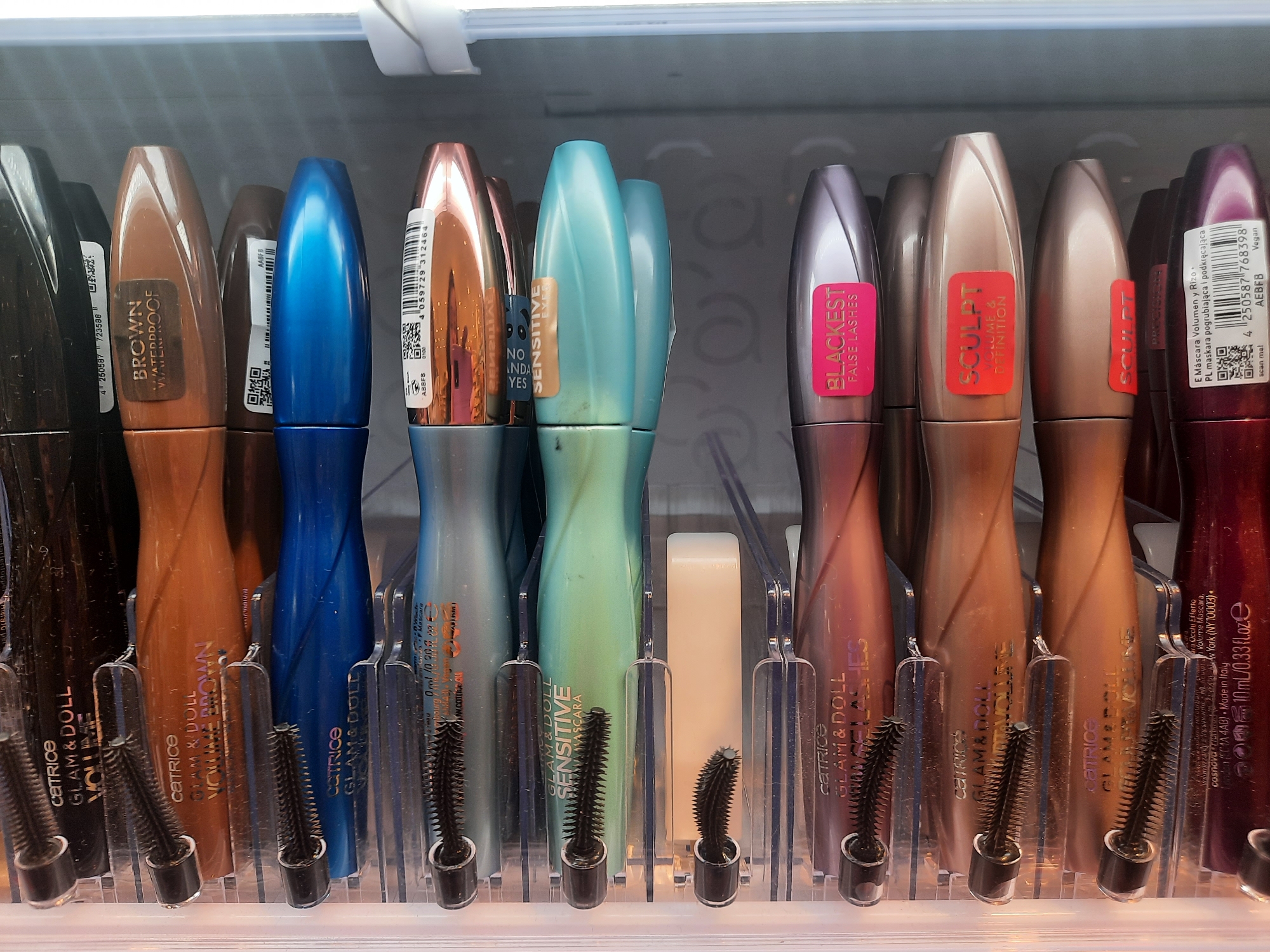 The image shows a close-up, eye-level view of a retail display featuring an assortment of mascara tubes. The products are arranged in clear plastic holders, with the mascara wands prominently displayed in front. The tubes vary in color, including brown, blue, teal, copper, and purple. Some tubes have labels indicating brands like "CATRICE," "GLAM & DOLL," "BROWN WATERPROOF," "SENSITIVE," "BLACKEST FALSE LASHES," and "SCULPT VOLUME & DEFINITION." Barcodes and product information are visible on some of the tubes. The display is well-lit, likely in an indoor retail environment. The context suggests a beauty products section of a store. The image was taken on the exterior facade of a store in Rosenheim, Germany, specifically in the vicinity of a "dm" drug store located at Ludwigsplatz 10, 83022 Rosenheim.