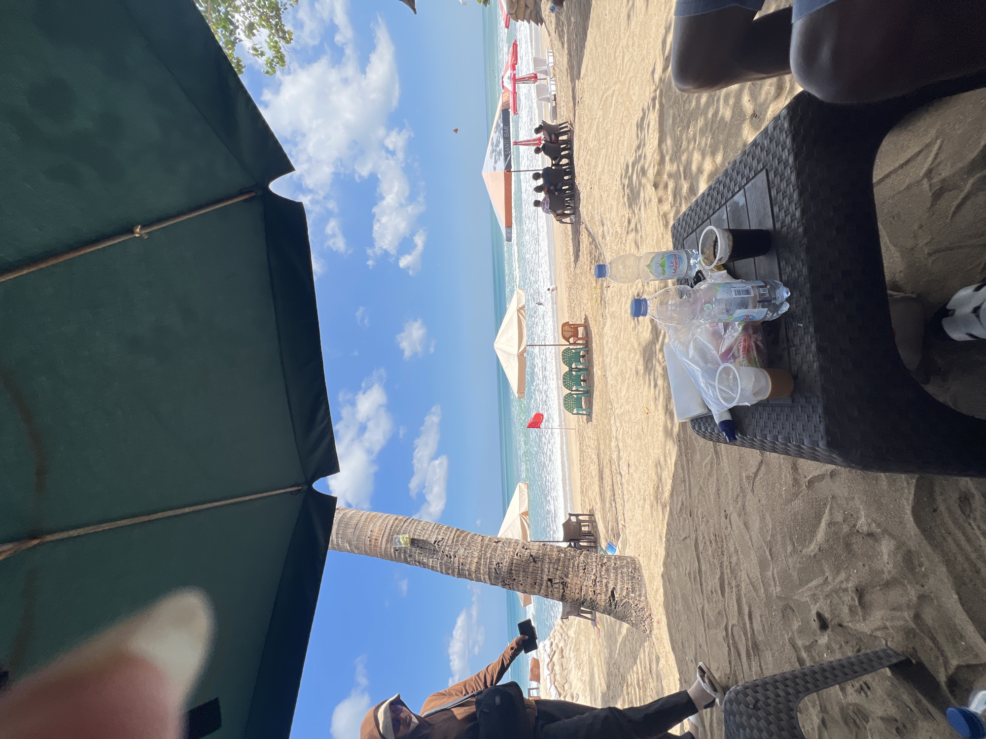 This image depicts a beach scene in Kuta, Indonesia, likely during daylight hours given the bright sun and blue sky. In the foreground, a dark-colored table sits on the sand, holding two plastic cups with beverages, two plastic water bottles, and a plastic bag. A person's legs, wearing blue shorts, are visible to the right of the table. The sand is textured and shows footprints.
To the left, a person wearing a hooded brown jacket, sunglasses, and a white hat is walking away from the camera. They are carrying a black bag over their shoulder and holding a dark object, possibly a phone, in their right hand. They are wearing dark pants and white flip-flops. A textured chair is visible to the far left.
In the mid-ground, a prominent palm tree stands on the sandy beach, with two clear plastic containers with what appears to be green fruit or decorations attached to it. Beyond the palm tree, the beach extends to the ocean, where gentle waves are breaking. Several beach umbrellas are scattered across the sand, some with chairs underneath. A red flag is planted in the sand near the water's edge. Further down the beach, a group of people are seated under an umbrella. The ocean stretches to the horizon under a bright blue sky with scattered white clouds. The scene suggests a relaxed atmosphere at a beach resort or cafe.