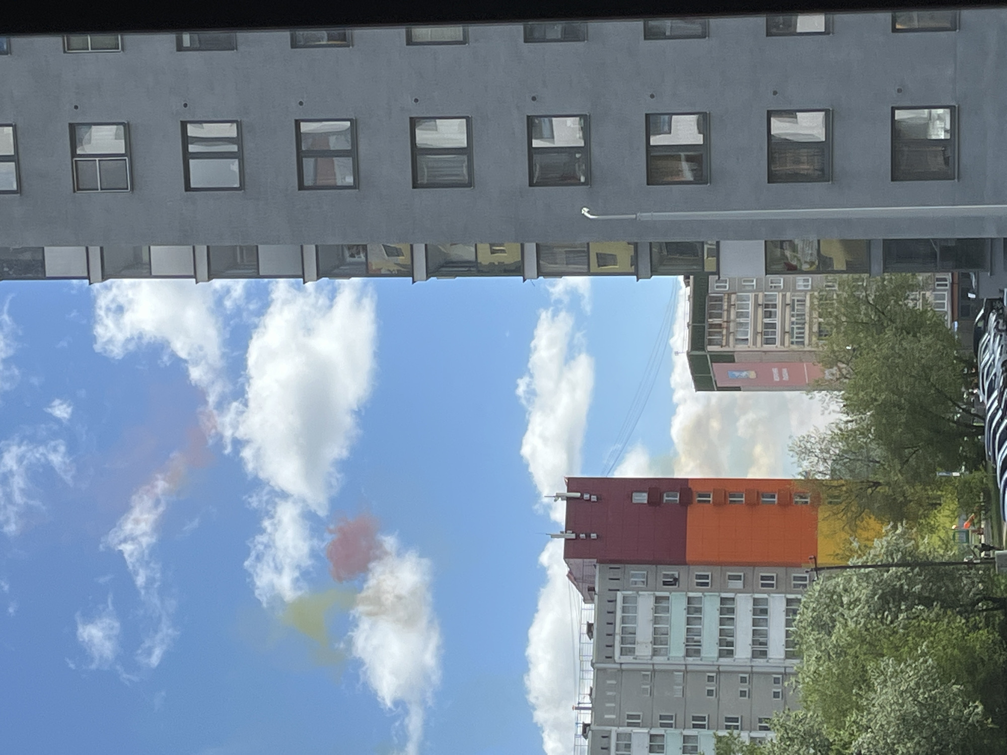 A worm's-eye-view photograph shows a street in Verkhnyaya Pyshma, Russia, featuring a clear blue sky with cumulus clouds positioned between two buildings. The buildings are located across from each other. The building at the top of the frame is gray with uniform square windows. The building below is a residential apartment building with a gray-and-white exterior and a colorful facade comprised of red, orange, and yellow sections. Adjacent to the second building are trees and street infrastructure. The photograph appears to be taken from a vehicle.