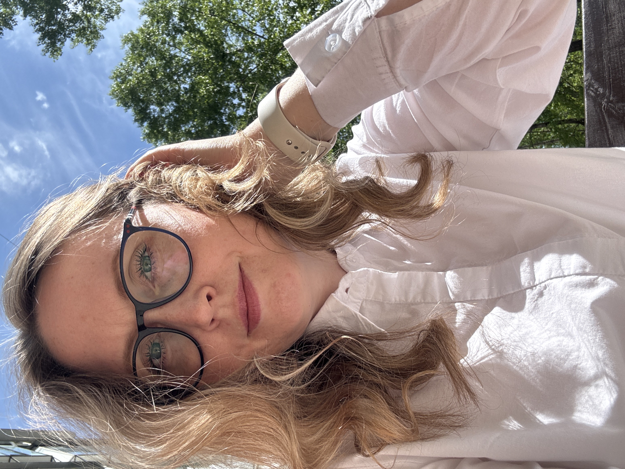 A woman with fair skin, blonde hair, and glasses is looking into the camera. Her right hand is touching the right side of her forehead. She is wearing a white, button-down shirt, and a light-colored wrist watch. The background shows trees, a blue sky, and a cloud-like pattern in the sky. It is not possible to determine from the image whether the location is Chaikovsky, Russia.