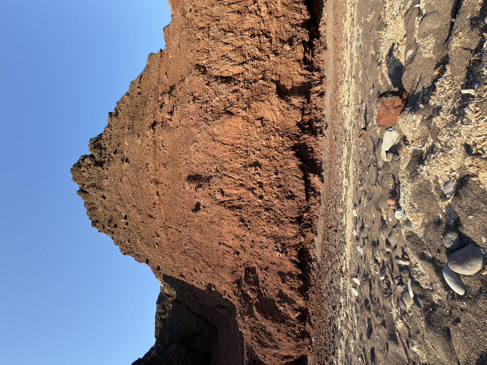 The image depicts a geological formation of reddish-brown rock situated on a beach under a clear blue sky. The rock face is textured and layered, with a prominent peak in the center, suggesting volcanic origin, common in the caldera of Thira Municipal Unit, Greece. The foreground is a beach composed of dark sand mixed with lighter debris and scattered pebbles and rocks of various sizes and colors. The lighting suggests it is either early morning or late afternoon, casting shadows on the beach and highlighting the textures of the rock. There are no people or animals visible in the scene.