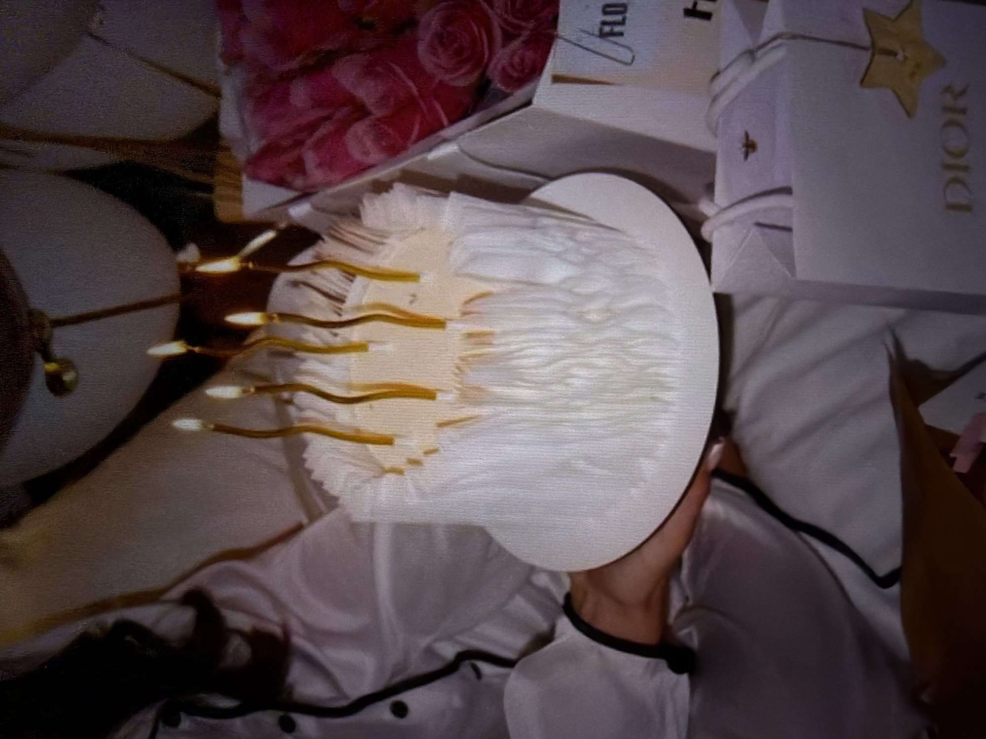 This media file shows a close-up view of a white cake decorated to resemble a jellyfish, with golden, strand-like candles extending from it. The cake is adorned with white ruffles or frosting that mimic the jellyfish's tentacles. A person's hand is visible holding the cake, with manicured fingernails suggesting attention to detail.
In the background, a white Dior shopping bag with a gold star accent and the "DIOR" logo is visible, along with a hint of pink roses, suggesting a gift or celebration context. The overall setting appears to be indoors, possibly a bedroom, given the soft lighting and the presence of satin pajamas worn by the person holding the cake. The scene conveys a sense of festivity and indulgence, likely a birthday or special occasion. No time of day or weather cues are discernible. The text "DIOR" is visible on the shopping bag, and "FLO" is partially visible on a white card or tag.