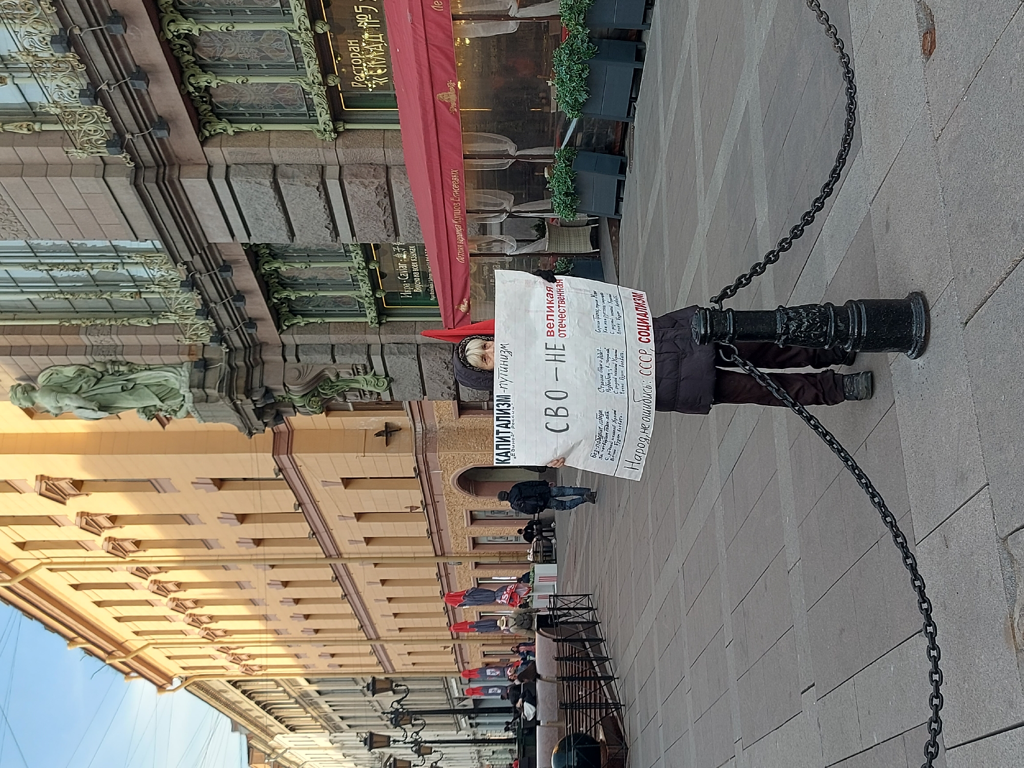 The image shows a person holding a sign in what appears to be Saint Petersburg, Russia. The location is indicated by the prominent "Restaurant" sign in Russian and the building design characteristic of the city. The person is standing near a black cannon, which is chained off. The sign they are holding has text in Russian, including the words "CAPITALISM-Putinism" (КАПИТАЛИЗМ-Путинизм), "Great Patriotic" (Великая отечественная), "SVO-NOT" (СВО-НЕ), "People, don't be mistaken: USSR, SOCIALISM" (Народ, не ошибись: СССР, СОЦИАЛИЗМ). The text on the sign includes several lines of poetry or prose. A few other people are visible walking in the background. There are tables and chairs set up suggesting an outdoor dining area or cafe along the building.