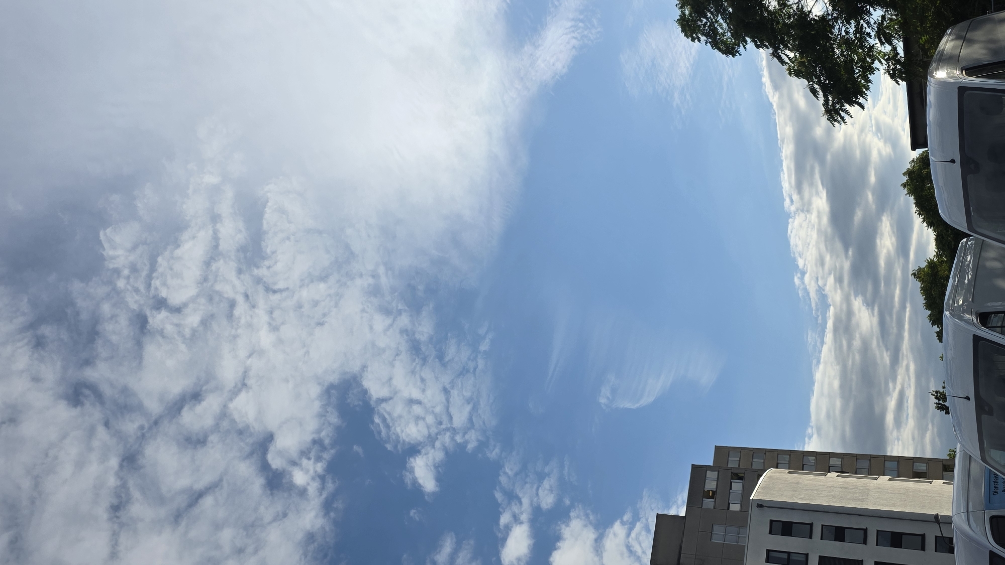 The image shows a daytime view of a cityscape with a bright blue sky filled with wispy white clouds. In the foreground, the tops of several white vans are visible, suggesting they are parked. To the left, two buildings stand side-by-side, one taller and gray with visible windows, and a shorter, lighter-colored building with a curved facade and dark-rimmed windows. Green trees line the bottom of the frame, partially obscuring the view of the vans and buildings. The lighting suggests it is either morning or late afternoon. The location is identified as Unknown, Unknown.