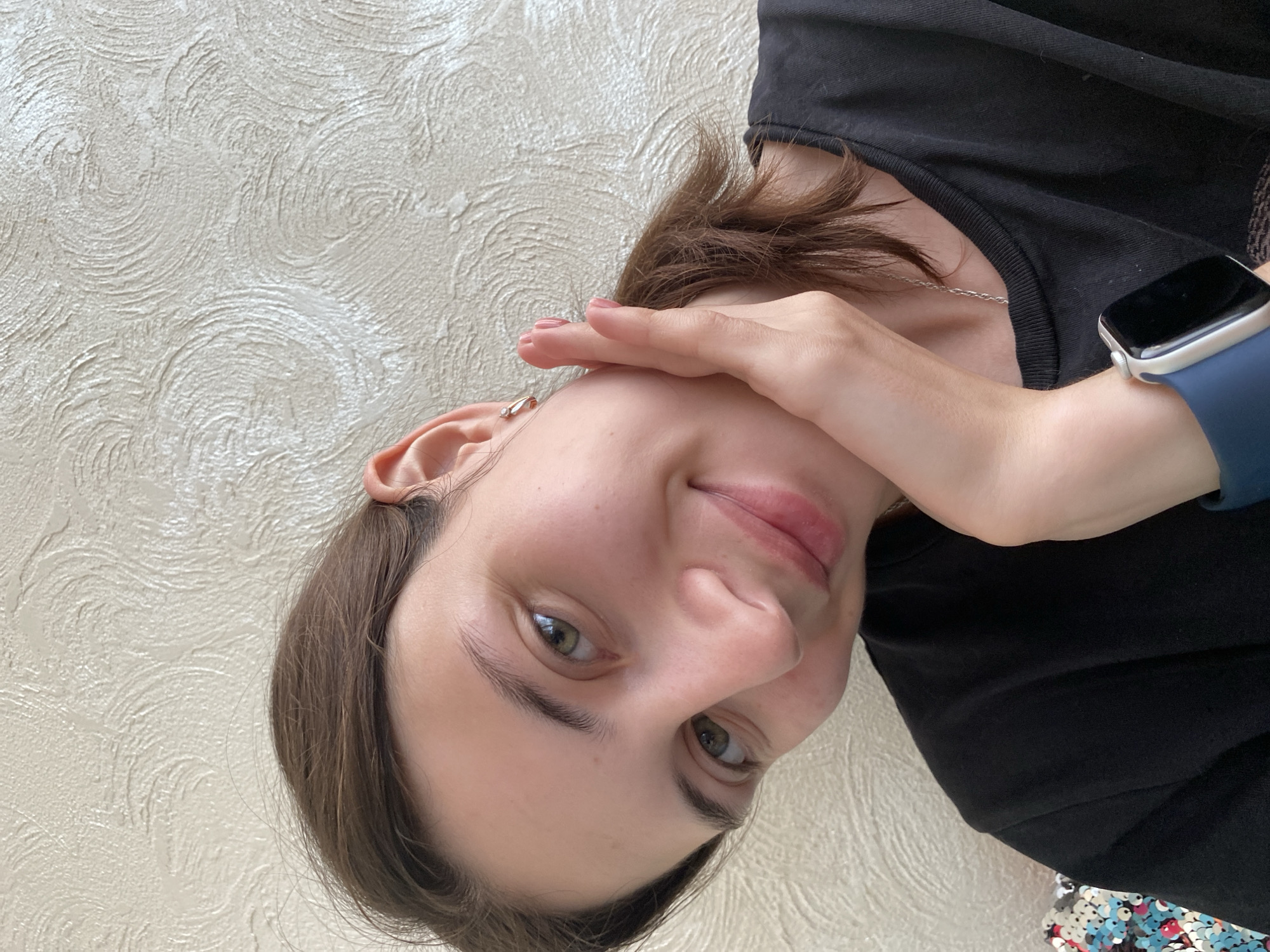 The media file is a close-up, vertically oriented selfie of a young person, likely female, against a textured cream-colored wall. The subject's head is tilted, and their face is partially obscured by their right hand, which is placed across their forehead and cheek. They are looking towards the camera with a gentle smile. Their eyes are green, and their lips are slightly parted. They have brown hair that is pulled back loosely, with some strands falling forward.
They are wearing a black t-shirt and a blue smartwatch on their left wrist. A thin silver necklace is visible around their neck. The lighting suggests daytime, possibly indoors given the uniform texture of the wall. The overall mood appears relaxed and intimate. There is no visible text in the image. The location context of Votkinsk, Russia, is provided as a hint but is not discernible from the image itself.