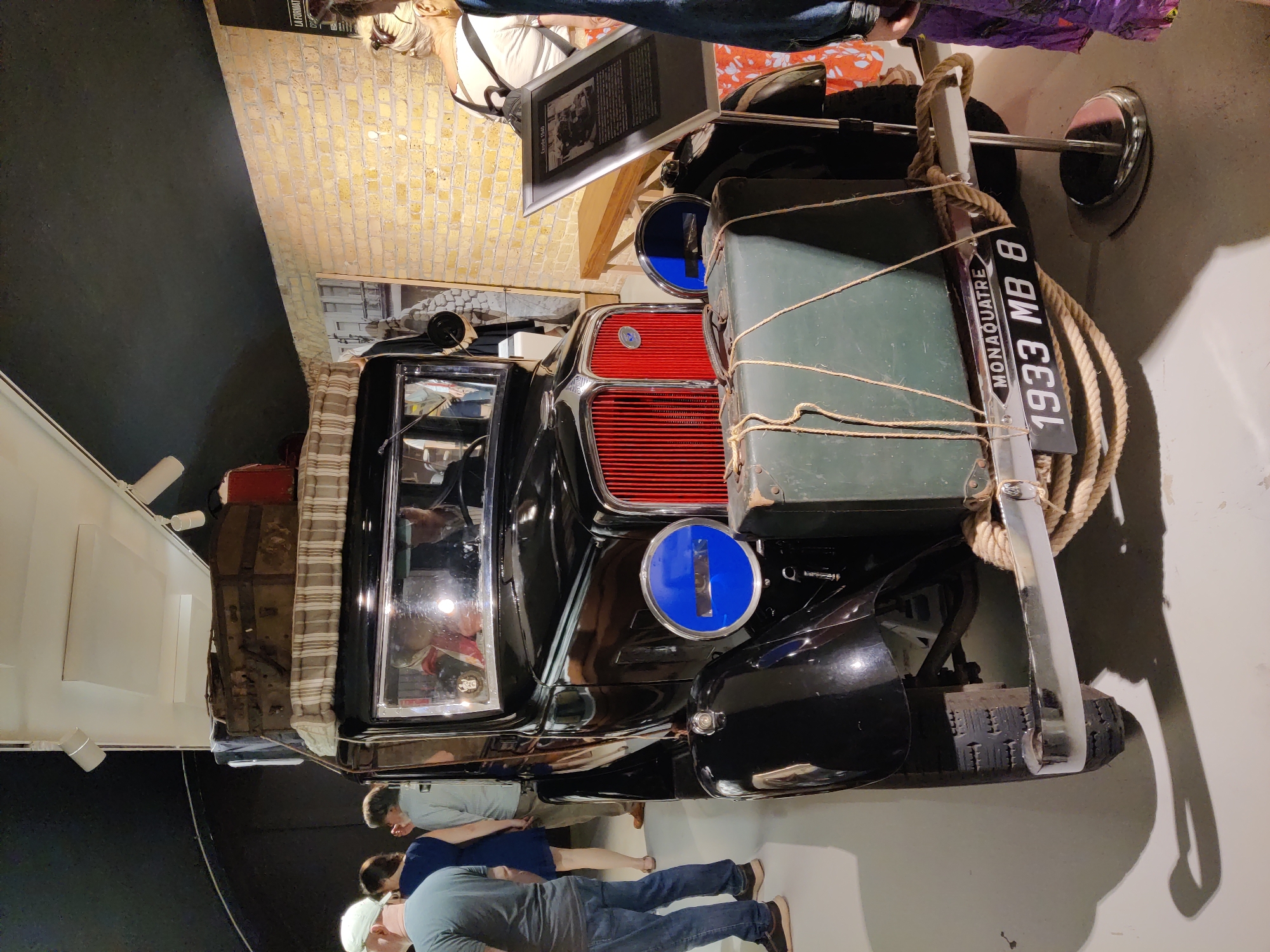 The image depicts a vintage black automobile, identified by a license plate reading "MONAQUATRE 1933 MB 8", prominently displayed indoors. The vehicle is loaded with luggage strapped to its roof and a large suitcase secured to its front bumper with rope. The car has a distinctive red grille and large blue circular headlights. Several individuals are visible in the background, appearing to observe the exhibit. The environment suggests an indoor exhibition or museum space, with a brick wall and lighting fixtures visible. The presence of the license plate and the "MONAQUATRE" inscription strongly suggests a connection to Dunkirk, France, as "Monaquatre" is a historical reference to the 1940 evacuation from Dunkirk, known as Operation Dynamo.