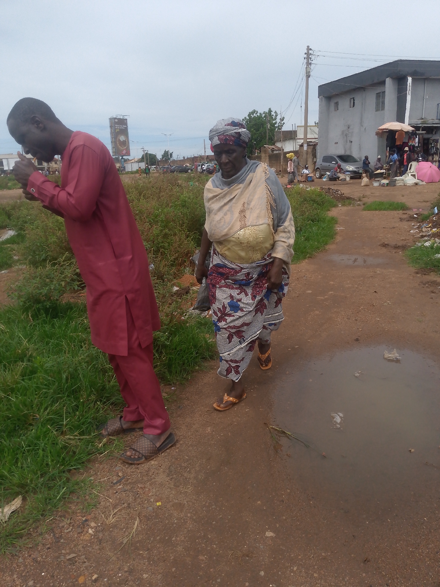 The image depicts an outdoor scene in what appears to be a market area or a roadside settlement in Bukuru, Nigeria. In the foreground, a man wearing a maroon outfit and patterned sandals is leaning down, appearing to light something, possibly a cigarette, with his thumb and forefinger. To his right, a woman wearing a headscarf, a beige shawl over a patterned dress, and flip-flops is walking away from the camera. Behind them, a dirt path with puddles of water is visible, leading towards buildings. In the middle ground, a two-story building with a dark roof and grey walls stands prominently, with several stalls and people gathered in front of it. Various vehicles, including cars and possibly a motorcycle, are present in the background. The environment appears to be somewhat cluttered, with some debris and vegetation visible. The sky is overcast, suggesting a cloudy or possibly rainy day.