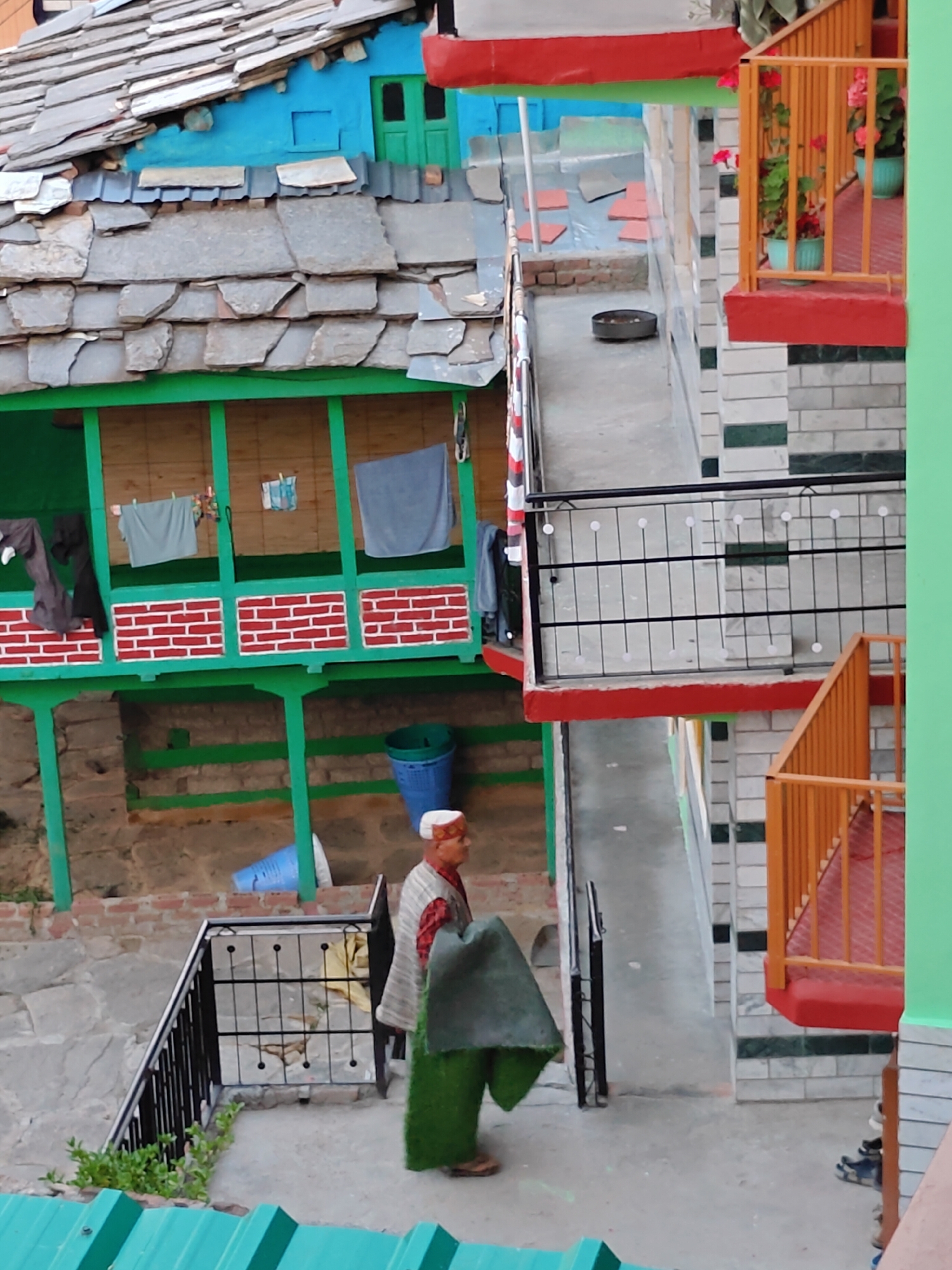 In Vashisht, India, an elderly man wearing a white and red patterned cap and a traditional shawl walks down a concrete stairway beside a multi-story building. The building features a combination of stone and modern brickwork, with brightly colored balconies and railings in red and orange. Above, a house with a blue facade has a traditional stone-tiled roof. Laundry hangs from a green-painted wooden balcony on a lower level. Blue and green plastic bins are visible near the base of the building. The scene depicts a residential area with distinct architectural styles.
