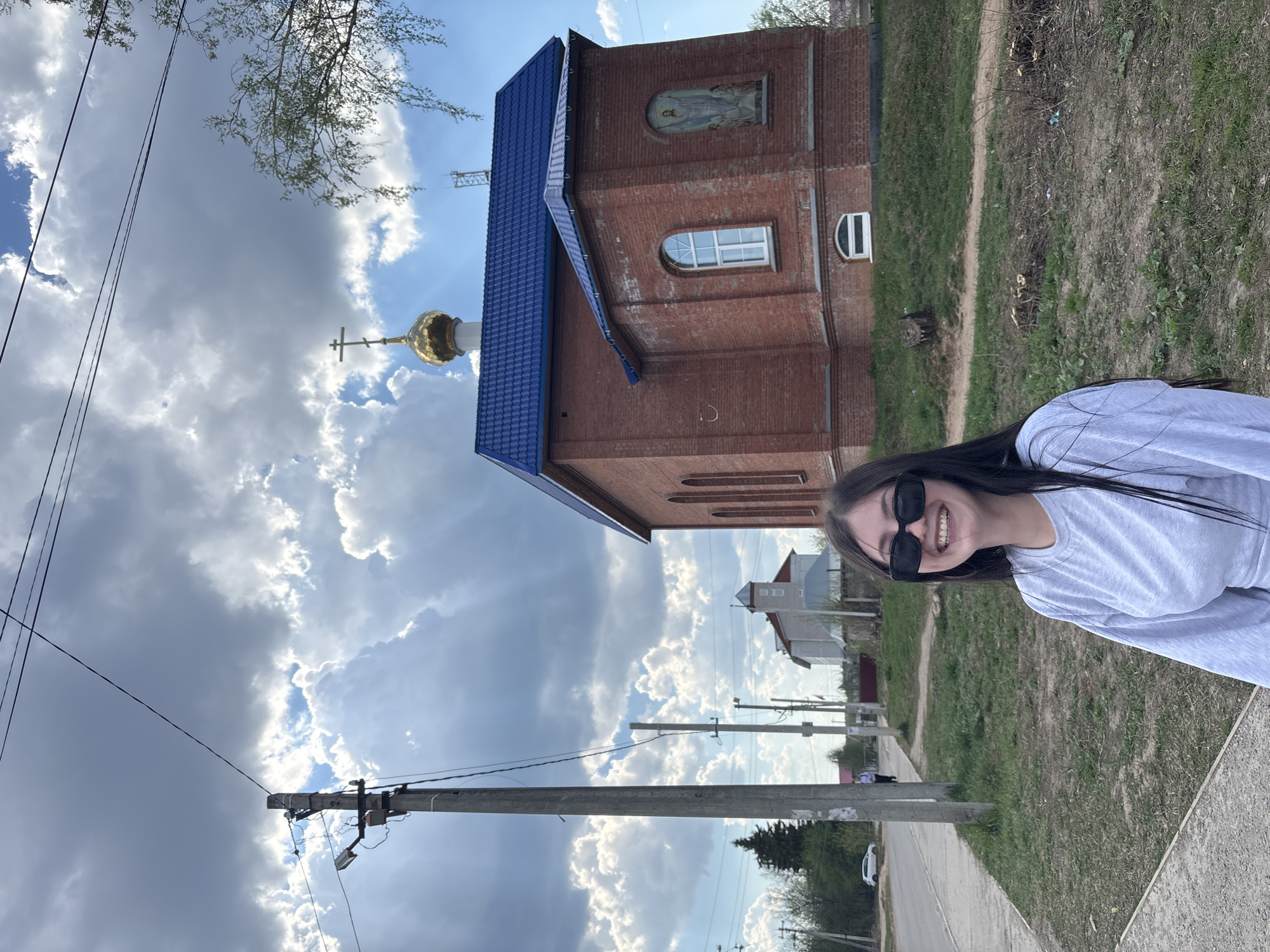 A young woman wearing sunglasses smiles at the camera. She stands on a sidewalk adjacent to a street. Power lines and a concrete utility pole run along the roadside. Beyond the street, there is a red brick building with a blue roof and a gold-domed structure with a cross atop it. The sky is mostly cloudy. Based on the metadata, the location is in Фоки, Russia.