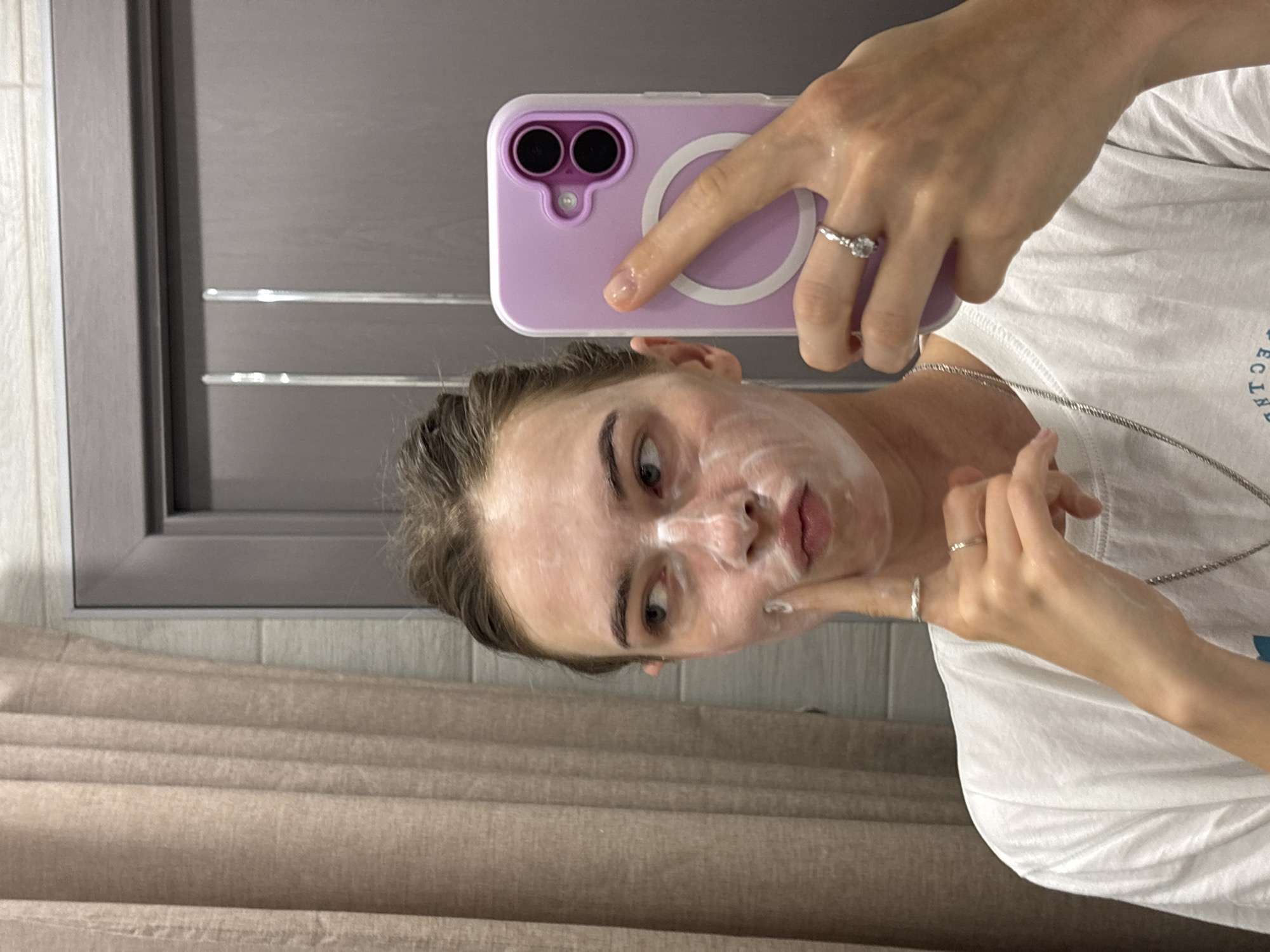 In a bathroom, a young woman with her hair pulled up takes a selfie in a mirror. She is applying a white face mask, with some of it smeared on her cheeks and nose. She is wearing a white t-shirt and multiple silver rings. Her phone, in a light purple case, is held up to take the photo. The background shows a beige curtain and a grey door. The location in the city of Perm, Russia is not discernible from the image.