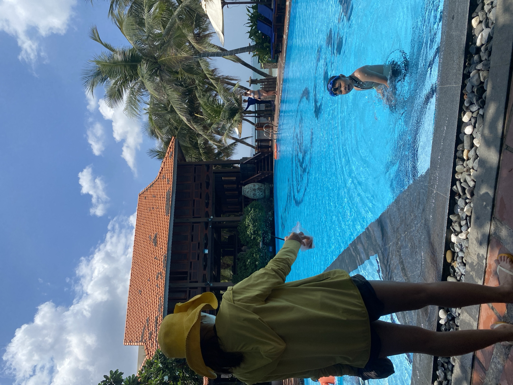 The image depicts a poolside scene in Phu Thuy Ward, Vietnam. In the foreground, a person wearing a yellow hat and a light green hooded jacket stands with their back to the camera, holding a clear plastic cup with a beverage. They are wearing dark shorts and flip-flops. In the middle ground, a young boy wearing blue swimming goggles is partially submerged in the bright blue swimming pool, looking towards the camera. The pool has a visible depth marker indicating 2 meters. Further in the background, a traditional Vietnamese-style building with an orange tiled roof and wooden shutters is visible, surrounded by palm trees and lush greenery. Several lounge chairs and umbrellas are set up near the pool. Other individuals can be seen in the background, some standing by the pool and others near the building. The sky is partly cloudy with a clear blue expanse. The poolside area is paved with tiles and bordered by small stones.