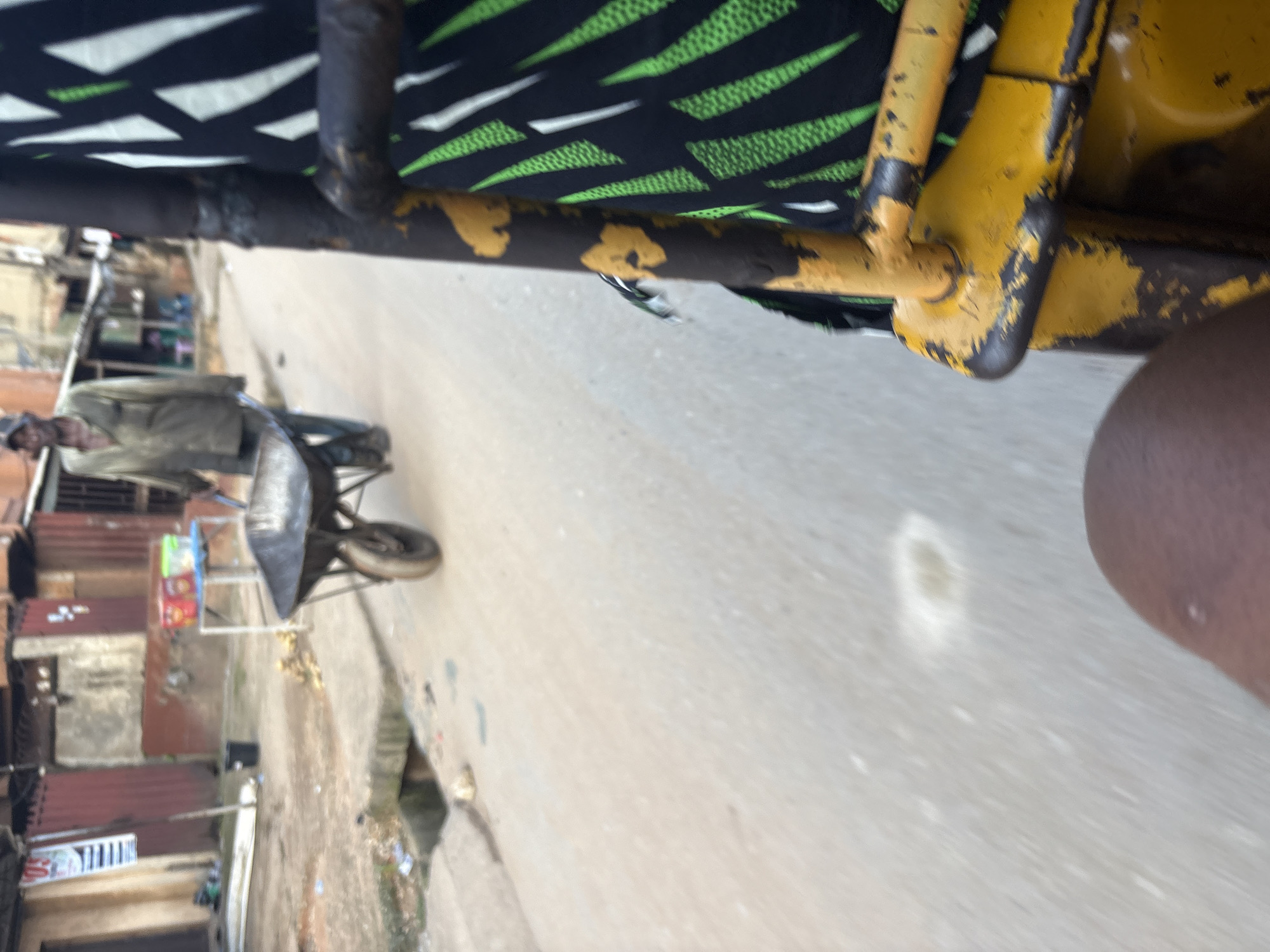 The image depicts a street scene in Onitsha, Nigeria. In the foreground, the yellow frame of a vehicle, likely a rickshaw or tricycle, is visible, along with a person's leg. In the middle ground, a man wearing a cap and a light-colored shirt is pushing a wheelbarrow along a paved road. The wheelbarrow appears to be partially loaded. In the background, there are buildings with corrugated metal siding, suggesting a commercial or residential area. Some items, possibly food containers, are displayed on a table near one of the buildings. A drain is visible alongside the road, and litter is scattered in the immediate vicinity. The overall impression is of a busy, urban environment.