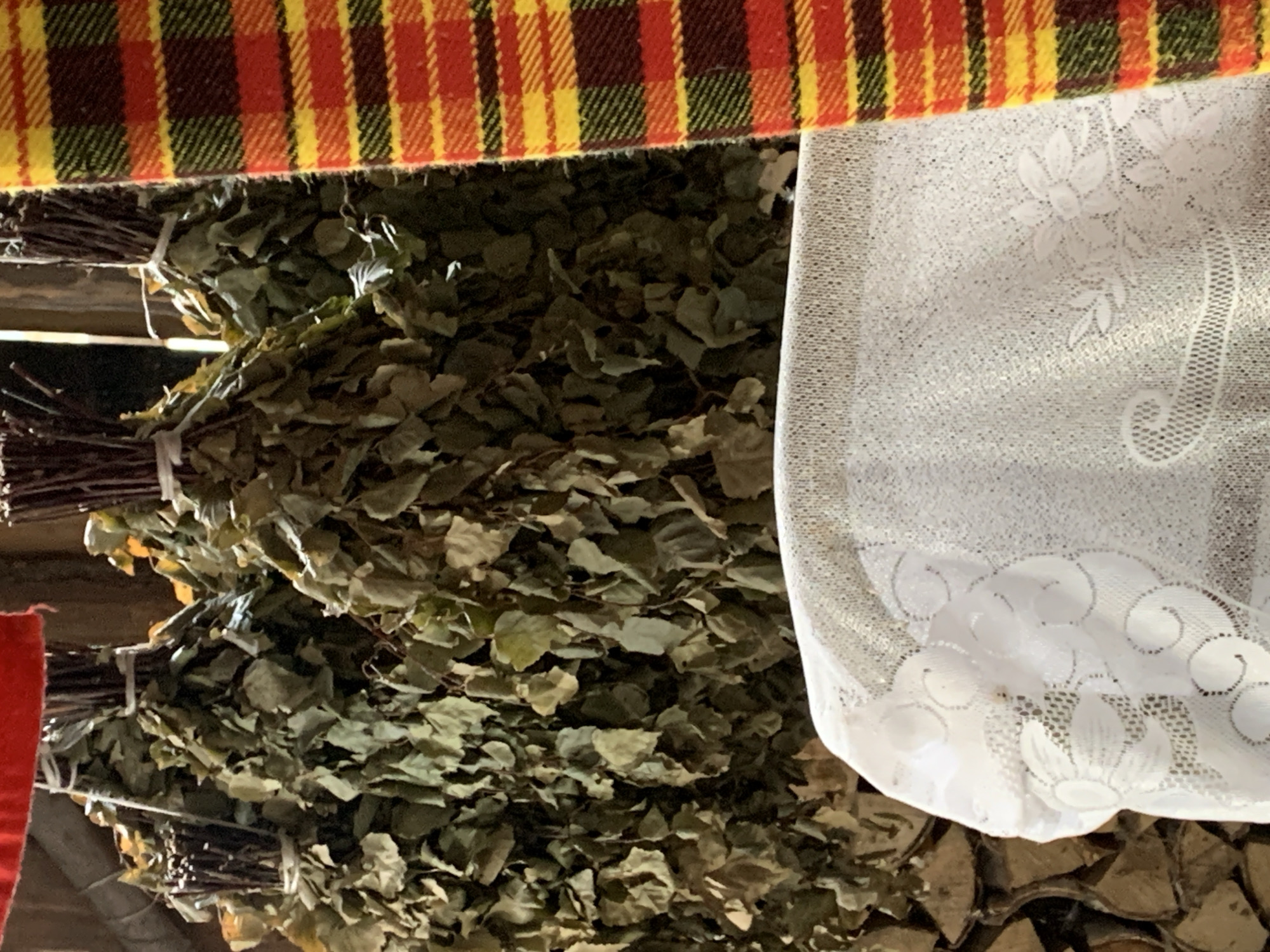 The image depicts an indoor scene with several bundles of dried foliage, likely birch brooms, hanging from a wooden structure. These bundles are tied with white twine and are densely packed with leaves and branches. To the right of the foliage, a strip of red, yellow, and green plaid fabric is visible. In the foreground, a white lace cloth covers an object, revealing a floral pattern. The background shows a stack of firewood and wooden beams, suggesting a rustic setting. The lighting appears dim, with some light filtering from above. The location is identified as Barsai, Russia.