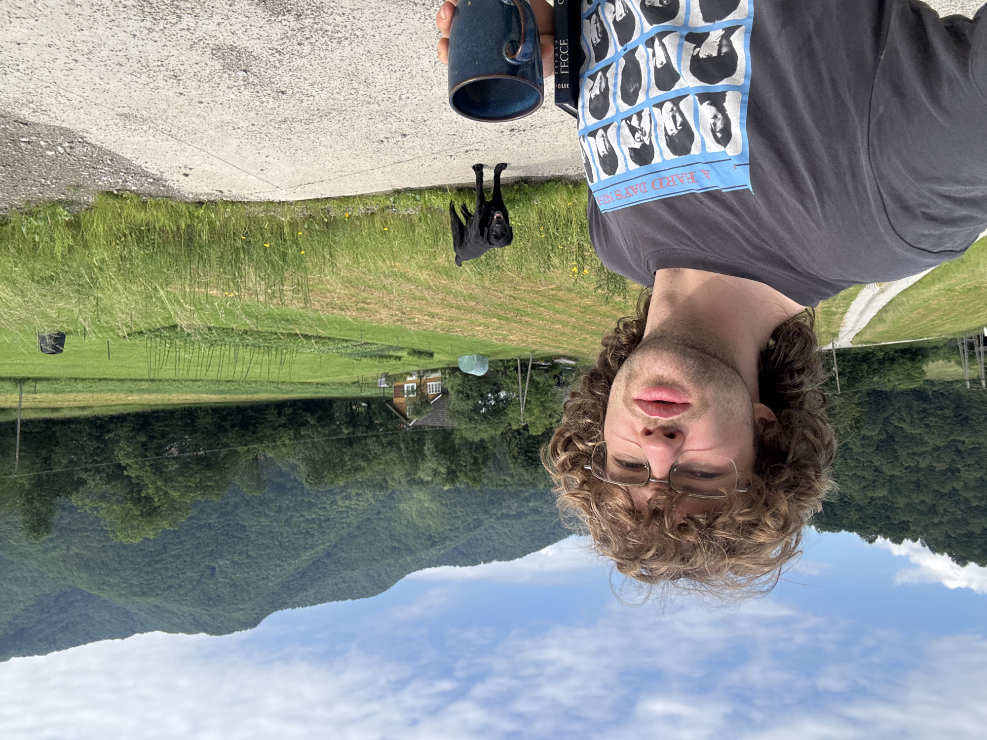 A young man with curly brown hair and glasses takes a selfie in front of a rural landscape. He is wearing a dark gray t-shirt with a print of The Beatles' album cover, "A Hard Day's Night." He holds a blue ceramic mug in his right hand and a black book in his left. Behind him, a black dog stands in the middle of a gravel path, looking towards the camera. The background features a green field, a small house with a red roof, and lush green hills under a partly cloudy sky. The location is described as Unknown, Unknown.