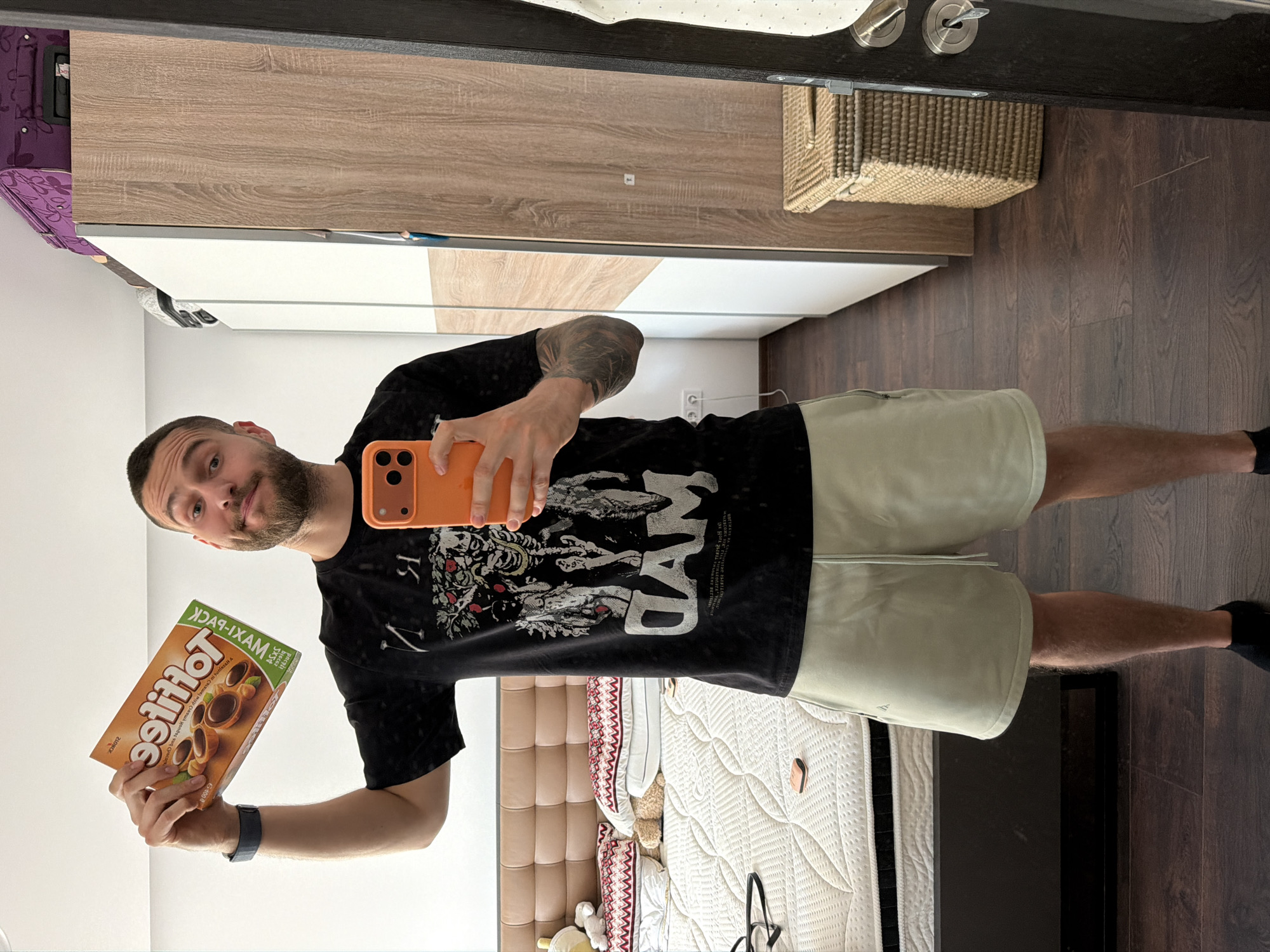 A bearded man with a forearm tattoo is taking a selfie in a mirror. He's holding a box of Toffifee candy in his left hand and a phone in his right hand. The reflection shows a bedroom with a bed, bedside table, a wardrobe with white and wood-grain doors, a woven basket, and a dark wood floor. While the city of Sofia is not identifiable from this image, the image is likely inside a residence.