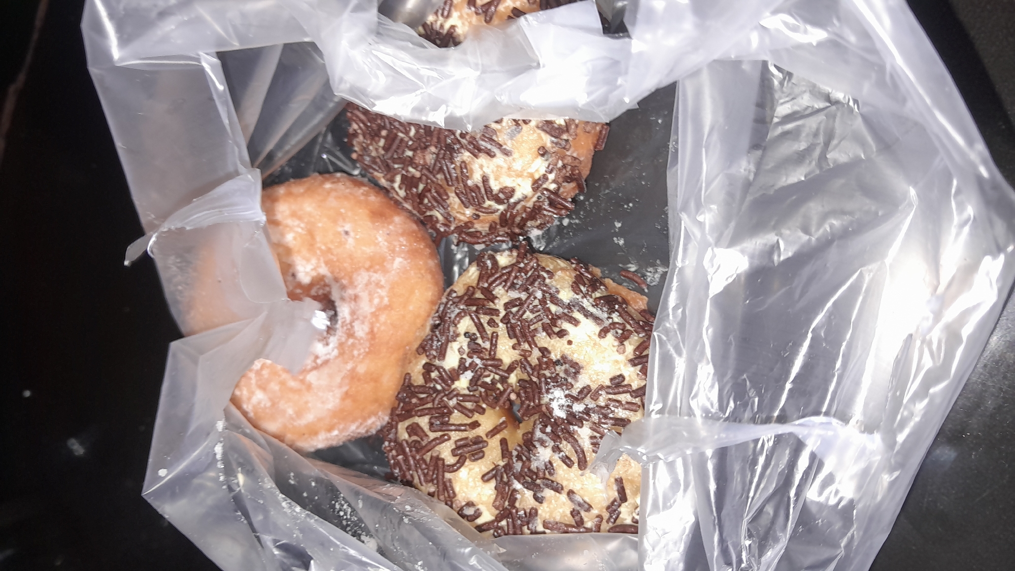 The image displays a clear plastic bag containing four donuts on a dark, flat surface. One donut, situated at the top center, is light brown, ring-shaped, and dusted with powdered sugar, exhibiting a partial consumption or tear on its upper edge. Two other donuts, positioned at the bottom left and bottom right, are light tan, ring-shaped, and heavily coated with dark brown chocolate sprinkles, also showing traces of powdered sugar. A fourth donut is partially visible behind the bottom right donut. The transparent plastic bag is crinkled, with light reflecting off its surface. No individuals or specific environmental markers beyond the dark surface are present. The scene is located in Lhokseumawe, Indonesia.