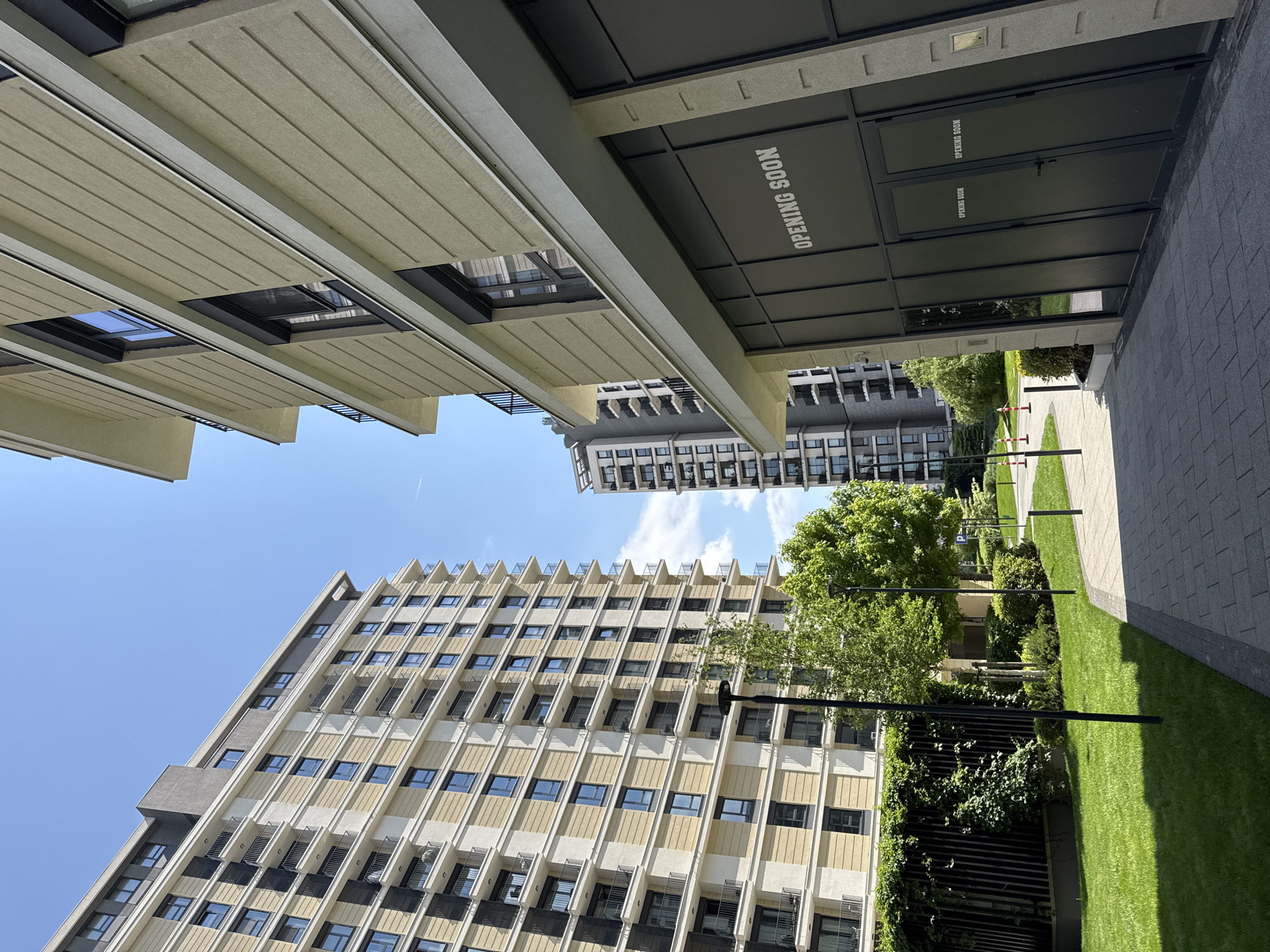 The image depicts a modern urban scene in Belgrade, Serbia. The camera is pointed upwards, capturing a view of several buildings and a landscaped courtyard. The buildings feature a blend of contemporary architectural styles, with a high-rise apartment complex visible with repetitive window patterns and light-colored facades. Underneath the building, there is an overhang with a sign that reads "OPENING SOON". The courtyard is well-maintained with green lawns, trees, and a paved walkway. The sky is clear and blue. There are no people present in the immediate foreground of the image.