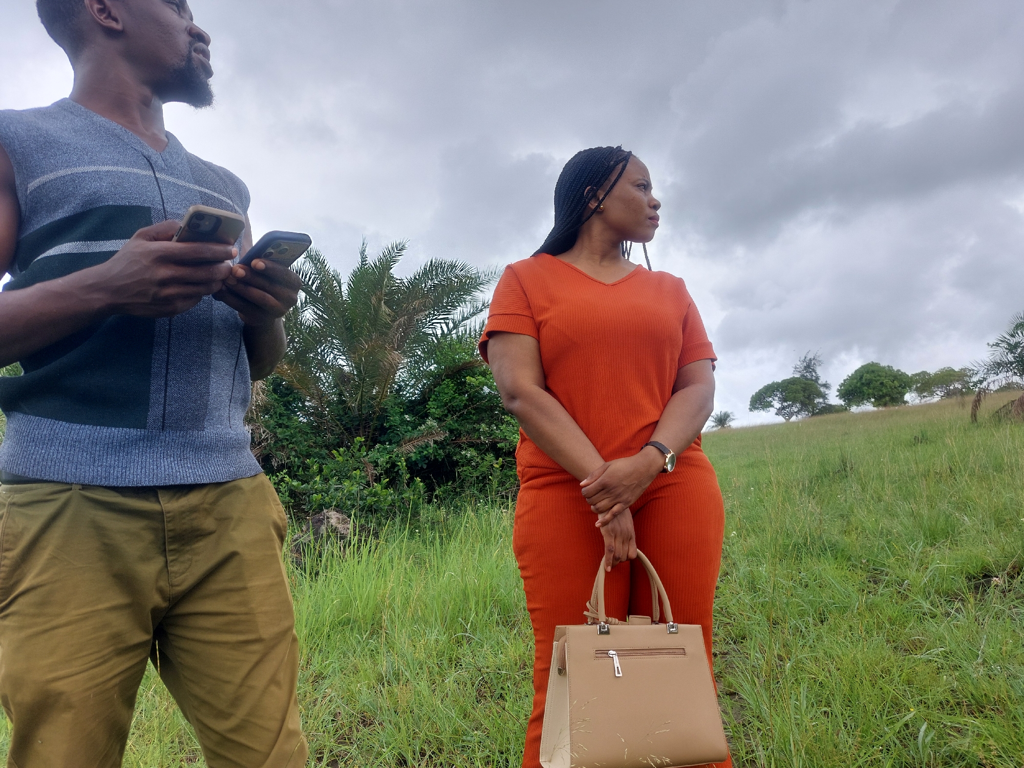 The image shows a man and a woman in a grassy field under an overcast sky. The man, positioned on the left, is dark-skinned, has a beard, and is wearing a sleeveless gray and black sweater over khaki pants. He is holding two smartphones in his hands. The woman, on the right, is wearing an orange pantsuit and holding a light brown handbag. She is facing to the right, away from the camera. The background consists of tall green grass, a few trees, and a grassy hill. Given the vegetation, the landscape appears to be in a rural area, therefore, I cannot tell the location in the city of Dar es-Salaam, Tanzania.