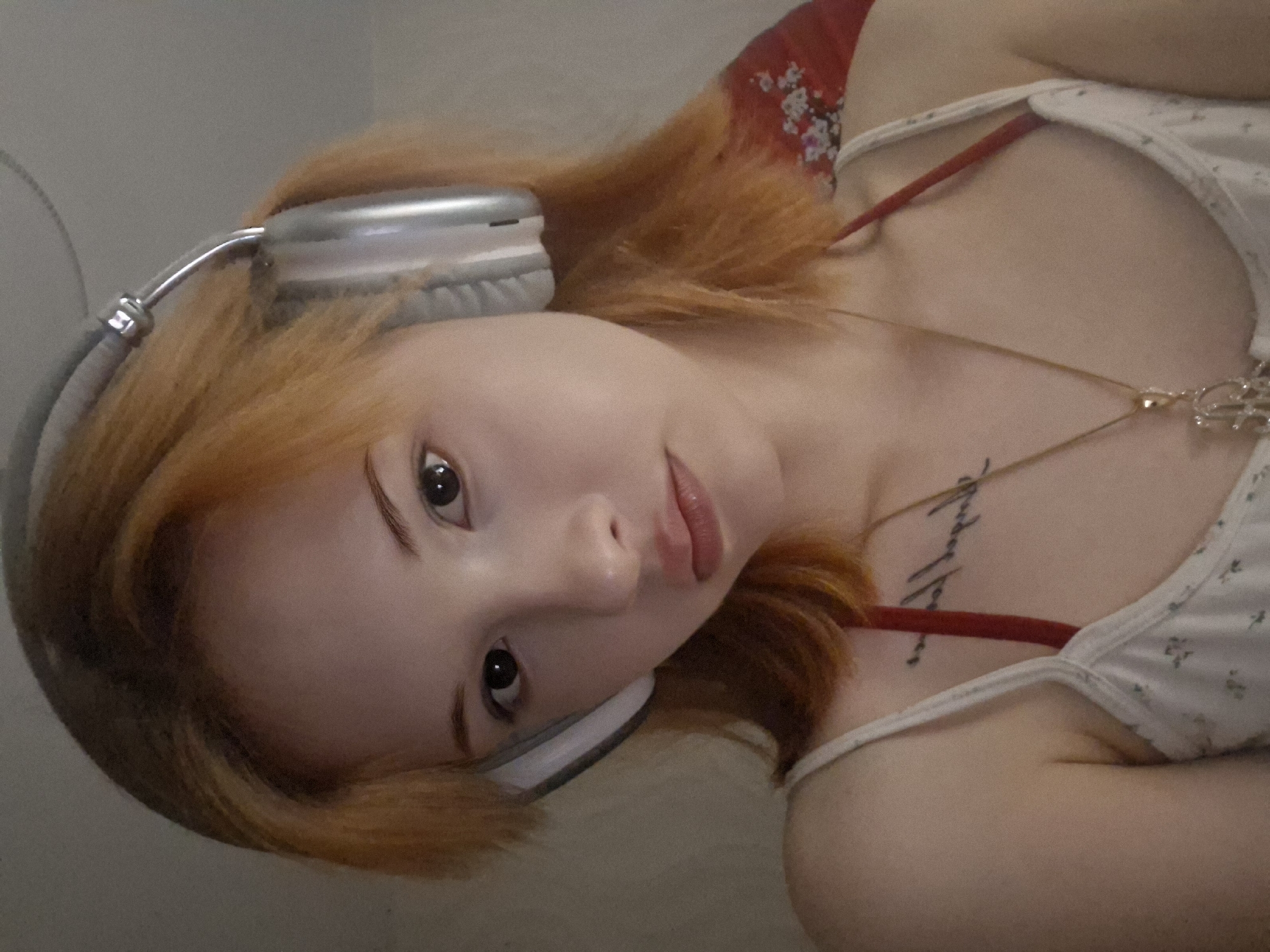 A young woman with reddish-blonde hair is lying down, wearing gray headphones and a white floral-patterned tank top with a red bra strap visible. She has a necklace and a tattoo on her chest. The background is indistinct, but there is a red floral pattern visible at the top of the frame. There is no information to determine if this photograph was taken in Chaikovsky, Russia.