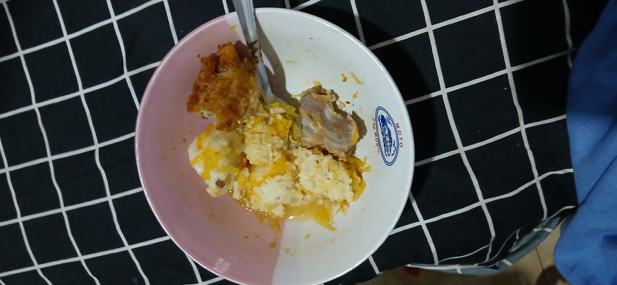 A close-up, overhead shot captures a serving of food in a pink and white bowl, placed on a black and white checkered surface. The food appears to be rice mixed with a yellowish sauce, possibly cheese or gravy, with some pieces of fried chicken and what looks like a chicken bone. A silver spoon is partially submerged in the food, suggesting it is being eaten. The bowl has a logo on its exterior, which appears to be a stylized image of a motorcycle with the text "Cap Mobil" and "MOTO" beneath it. The checkered surface extends to the edges of the frame, with some blue fabric visible at the bottom. There is no discernible indication of a specific location within Cigombong, Indonesia, beyond it being a scene depicting a meal.