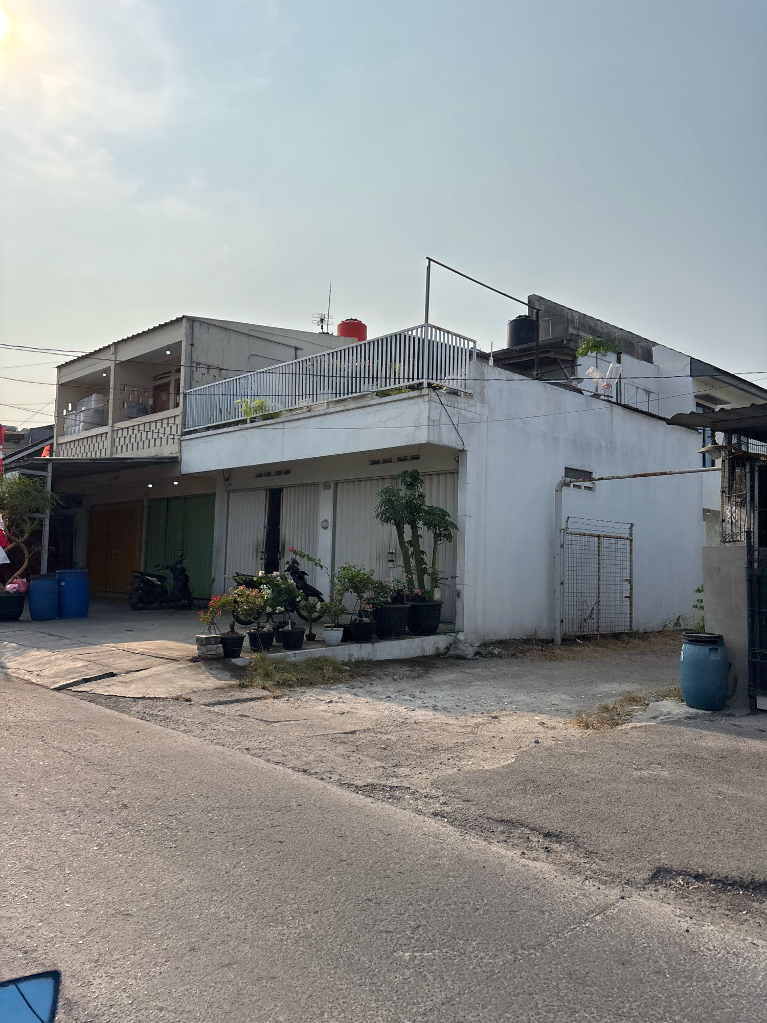The image shows a street scene in Bekasi, Indonesia, with a two-story building featuring a balcony on the upper floor and multiple roller doors on the ground level. The building is predominantly white with some areas of exposed concrete. Several potted plants are arranged in front of the building, and two blue plastic barrels are visible to the left. Two motorcycles are parked near the roller doors. The sky is clear with some sunlight, indicating it is daytime. The road in the foreground appears to be asphalt and shows some signs of wear and tear. A metal gate with wire mesh is seen to the right of the building. A red water tank is visible on the roof of the building, along with a black water tank and some other rooftop structures.