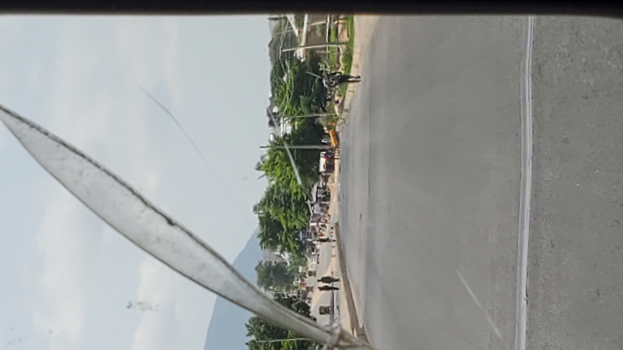 A street scene in Jalingo, Nigeria, is captured through a cracked windshield. The road is paved and appears to be a main thoroughfare with some traffic and pedestrian activity. On the left side of the road, several people are standing near parked vehicles, including what appears to be a tricycle taxi. Further down the road, there are more people, market stalls, and trees lining the street. On the right side of the road, a person is walking away from the camera, carrying a long object over their shoulder. In the background, there are buildings and a mountain or large hill. The sky is overcast. The overall impression is of a busy, typical day in a Nigerian city.