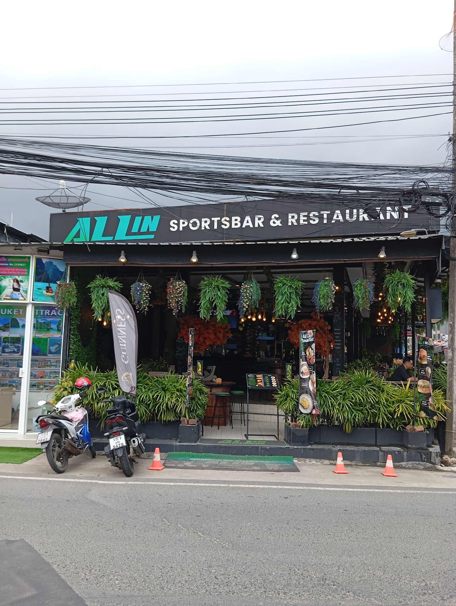 The image shows the exterior of an establishment in Karon, Thailand, identified by the sign "ALL IN SPORTSBAR & RESTAURANT". The facade features a black awning with the establishment's name in white lettering. Hanging plants and artificial foliage decorate the entrance, creating a lush, green atmosphere. Several tables and chairs are visible inside, suggesting an indoor seating area.
Outside the establishment, two motorcycles are parked on the pavement. The motorcycle on the left is white and blue, while the one on the right is black. In front of the entrance, there are several orange traffic cones placed on the pavement.
A man is seated at one of the tables inside the establishment, appearing to be a customer. Another person, potentially an employee, is standing near the right side of the entrance.
The street in front of the establishment is paved, with a white line marking the lane. The sky is overcast, indicating a cloudy day. The overall scene depicts a typical street view of a commercial establishment in a tourist area.