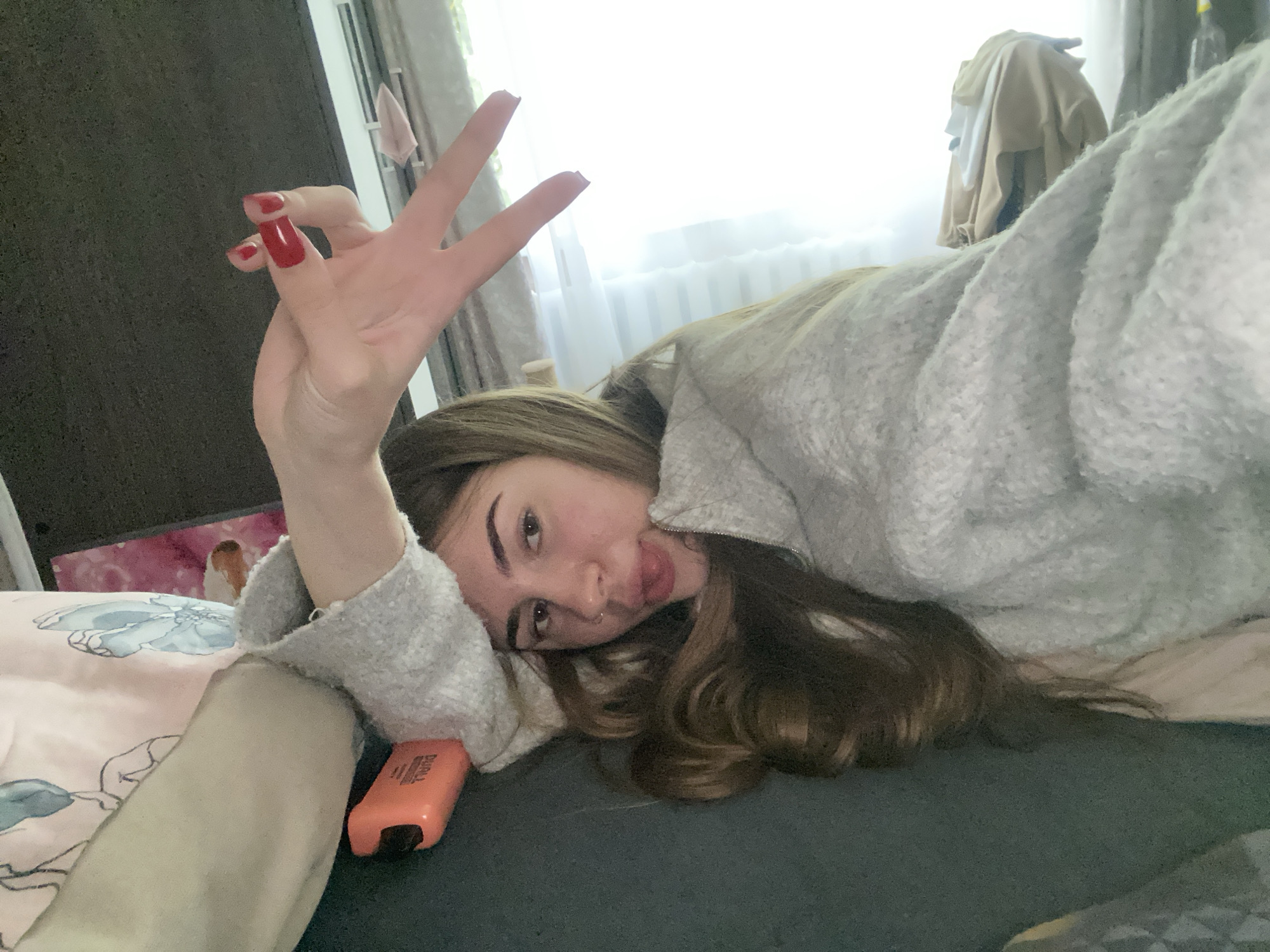 A young woman is depicted in a casual indoor setting, likely a bedroom. She is lying down, resting her head on a soft, light grey, fuzzy garment or blanket. Her long, brown hair is spread around her. She looks towards the camera with a playful expression, a small portion of her tongue visible, and a stud piercing in her left nostril.
Her left hand is raised, making a peace sign with two fingers extended. Her nails are long, rectangular, and painted a vibrant red. Beneath her left arm, a pink pillow with faint blue floral patterns is visible. An orange object, possibly a highlighter or a small device, lies near her arm; faint, partially legible text on it might read "DURLA" or similar.
The background includes a brightly lit window with white curtains on the right, indicating daytime. Further back, clothes appear to be draped over furniture or a hanger. A dark wall or furniture piece is visible on the far left. The overall impression is relaxed and informal, capturing a moment of ease.