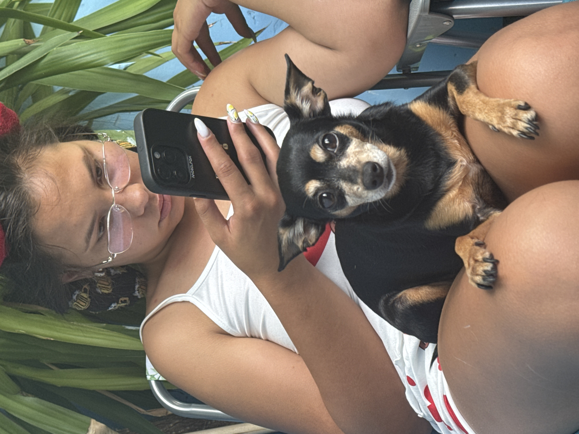 The image depicts a close-up of a young woman sitting outdoors, holding a smartphone and a small black and tan dog. The woman is wearing a white tank top with a red heart design and white shorts with red heart patterns. She has pink-tinted glasses and her hair is styled with red accessories. The dog is positioned on her lap, looking directly at the camera with its ears perked. The background includes lush green foliage and a blue wall. The overall impression is a casual, candid moment captured in natural light. There are no identifiable landmarks or specific details that would indicate a location within the city of Sofia, Bulgaria.