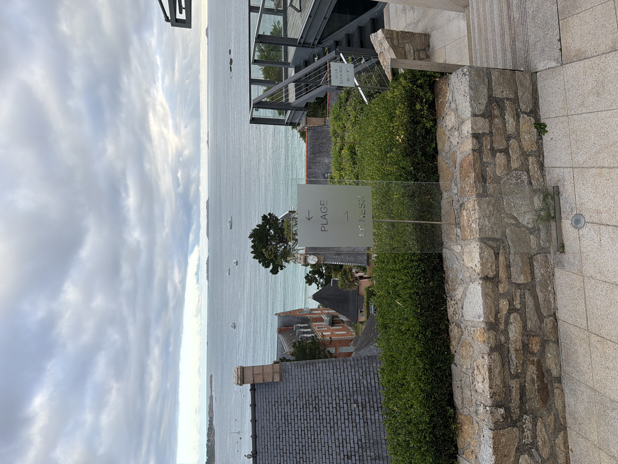 An overcast day in La Clarté, France, offers a view of the sea with several boats dotted on the water. In the foreground, a glass sign with an arrow pointing left and the word "PLAGE" (beach) is positioned in front of a stone wall and lush green bushes. To the right of the sign, another smaller sign indicates "VILLA EDEN ROC" with arrows and numbers. A modern metal staircase with glass railings leads up to a deck area on the right side of the frame. Behind the bushes, residential buildings with slate roofs and red-brick facades are visible, nestled amongst trees. The overall scene depicts a coastal landscape with a mix of natural beauty and modern architecture.