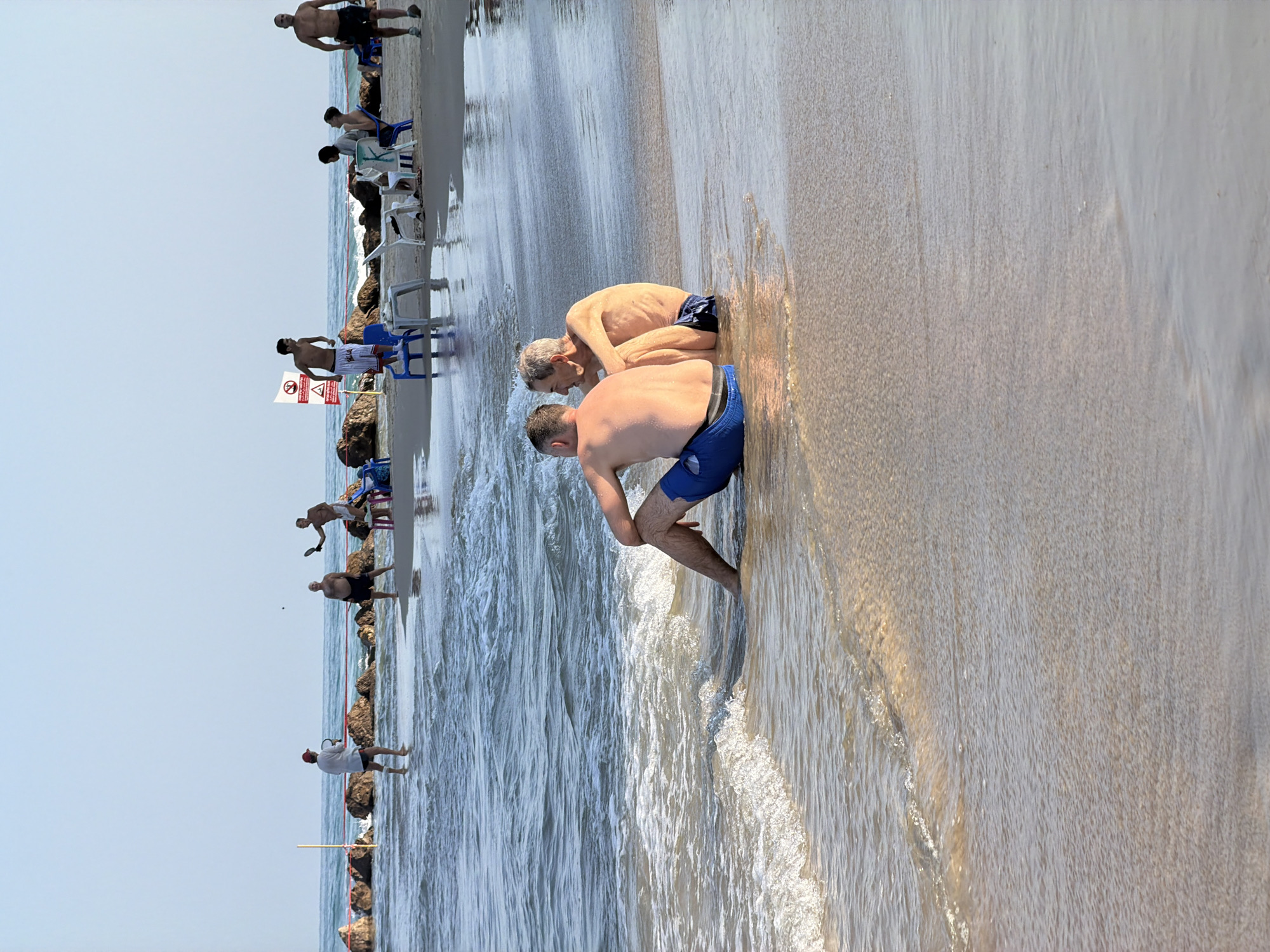 Two men are sitting on the beach in Bat Yam, Israel, with their backs to the camera, facing the sea. They are wearing swimwear, and one of them is sitting closer to the water, with the waves gently lapping around his legs. In the background, several other people are visible on the beach and in the water. Some are playing a game that appears to be paddleball, while others are sitting on chairs. The scene is bathed in sunlight, indicating it is daytime. The environment is a sandy beach with a calm sea.