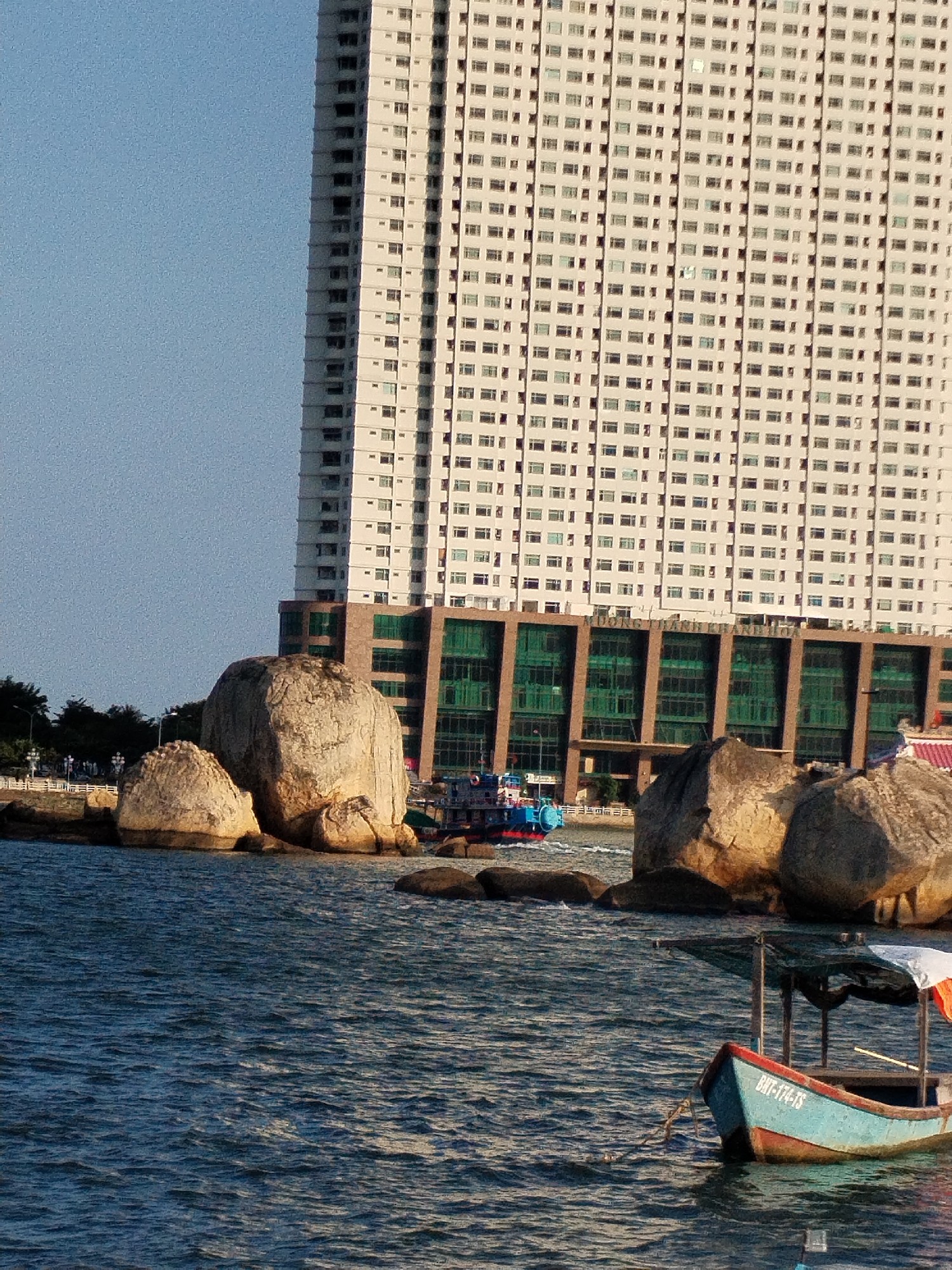 The image shows a waterfront scene in Bac Nha Trang Ward, Vietnam, with a tall, modern building dominating the background. The building has a light-colored facade and numerous windows arranged in a grid pattern. At its base is a structure with dark green glass windows.
In the middle ground, there are several large, rounded rocks along the shoreline. A blue and red boat is navigating the water near these rocks.
In the foreground, a small, blue boat with a partially covered awning is moored. The boat is labeled "BNT-174-TS." The water is dark blue and rippled. The sky is a clear, light blue.